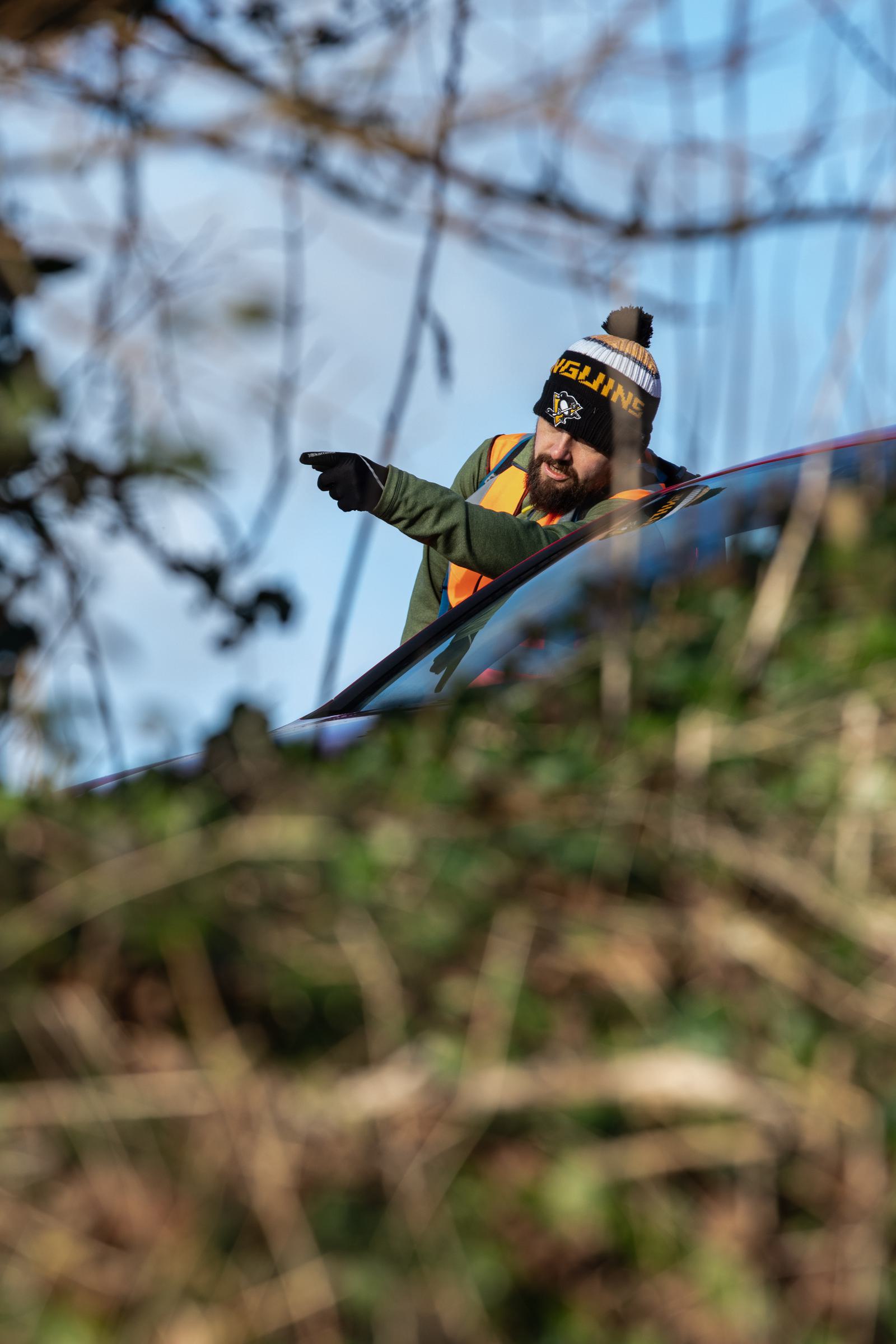 A person wearing a winter hat with a pom-pom, glasses, and a safety vest is pointing towards something while partially obscured by branches and foliage. They appear to be near a vehicle and are gesturing intently. The background shows a clear, sunny sky.