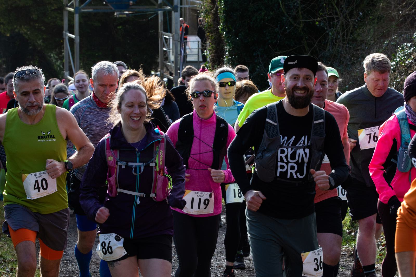 A group of runners in colorful athletic gear participating in a race on a wooded trail. Some wear bib numbers and hydration packs. They are surrounded by lush greenery, with a clear, sunny sky above. The atmosphere appears energetic and focused.