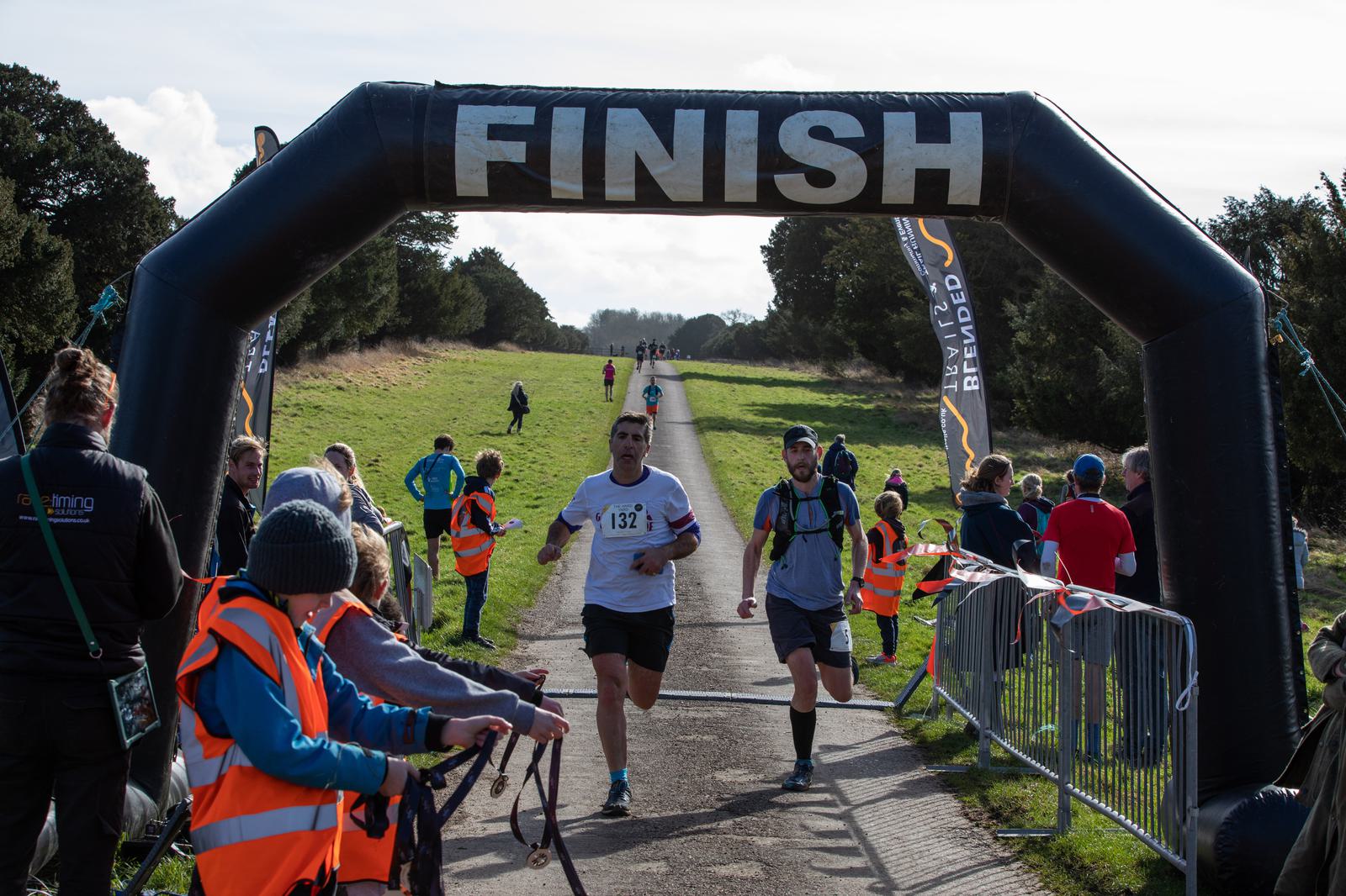 Runners are crossing the finish line of a race, passing under a large black inflatable arch with "FINISH" written on it. Spectators and event staff, some in orange vests, are present along the sides, and the background shows a path lined with greenery.