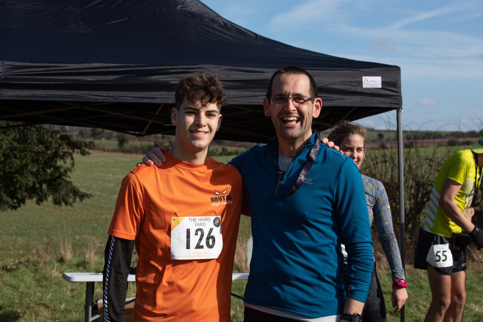 Two men standing under a black canopy tent at an outdoor event. The man on the left wears an orange shirt with a race number (126) and smiles. The man on the right wears glasses and a blue shirt, also smiling. Other participants and fields are visible in the background.