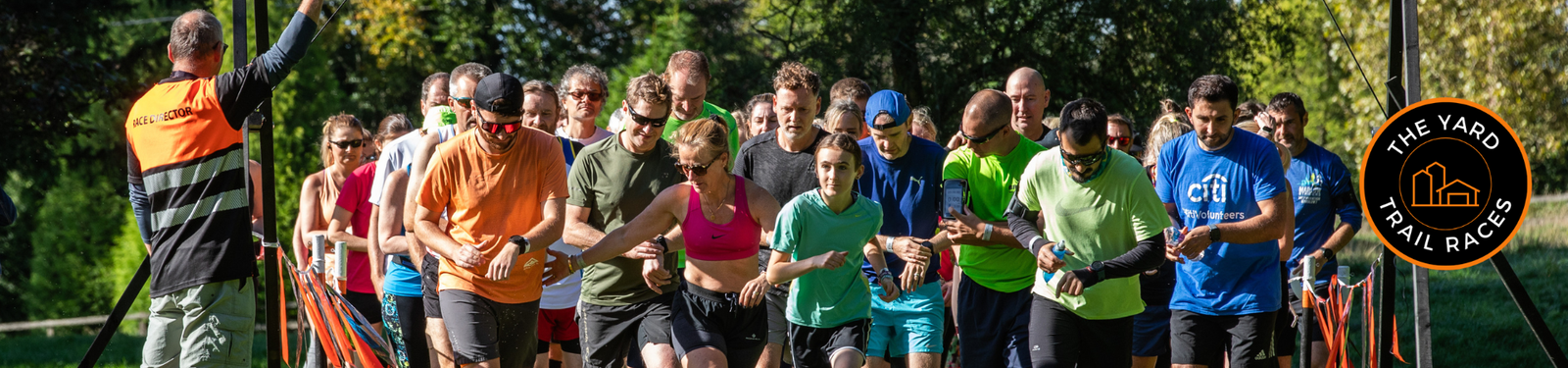 A group of runners in colorful athletic wear start a trail race in a green park, guided by an official in an orange vest. "The Yard Trail Races" logo appears on the right side of the image.