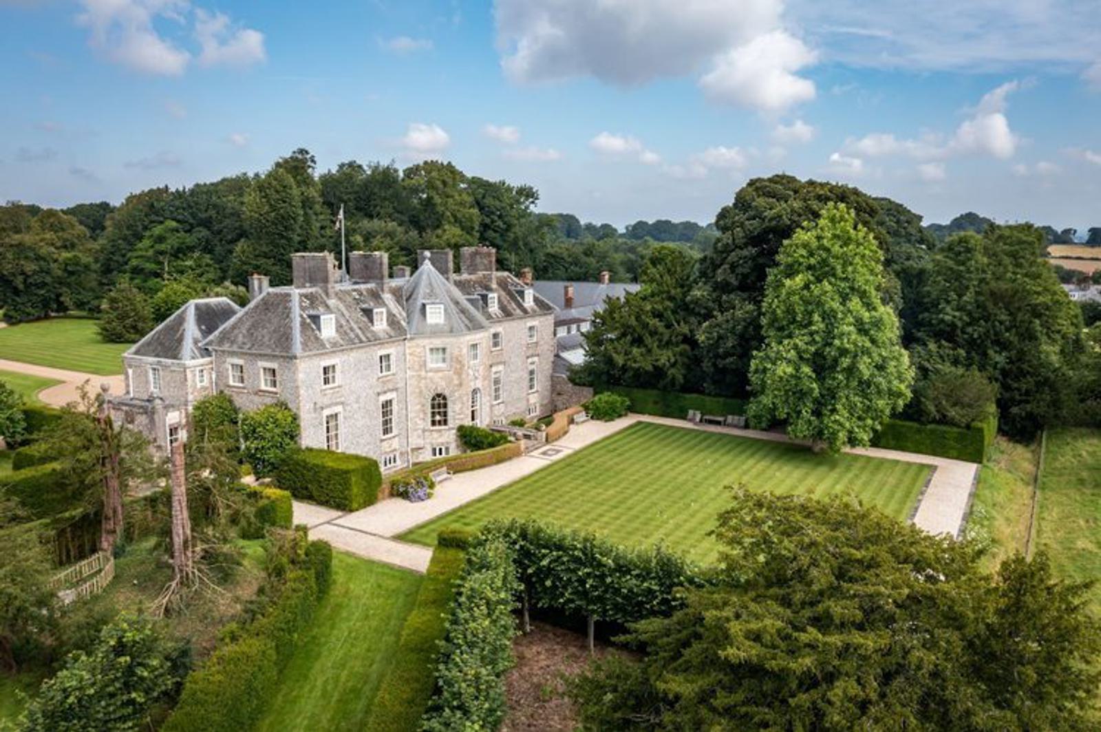 Aerial view of a large, historic stone mansion surrounded by lush gardens and trees. The building has a slate roof and multiple chimneys. Well-manicured lawns and hedges are visible, creating a serene and picturesque landscape.