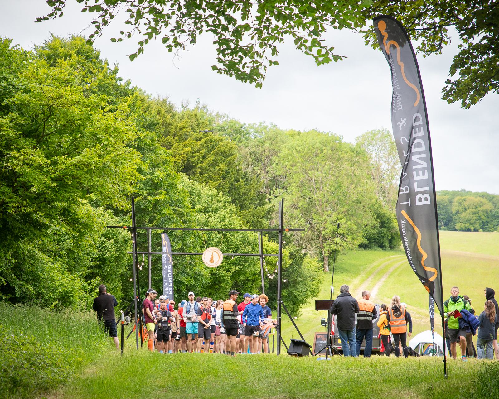 A group of runners eagerly waits at a starting line in a lush, green outdoor setting. Two vertical banners flank the start area, and the path ahead leads through scenic countryside. Spectators and event staff are present.