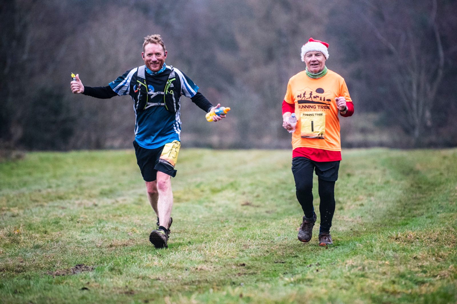 Two men are running on a grassy trail; one wears a blue athletic outfit, the other wears an orange shirt and a Santa hat. Both are smiling and appear to be participating in an outdoor event or fun run.