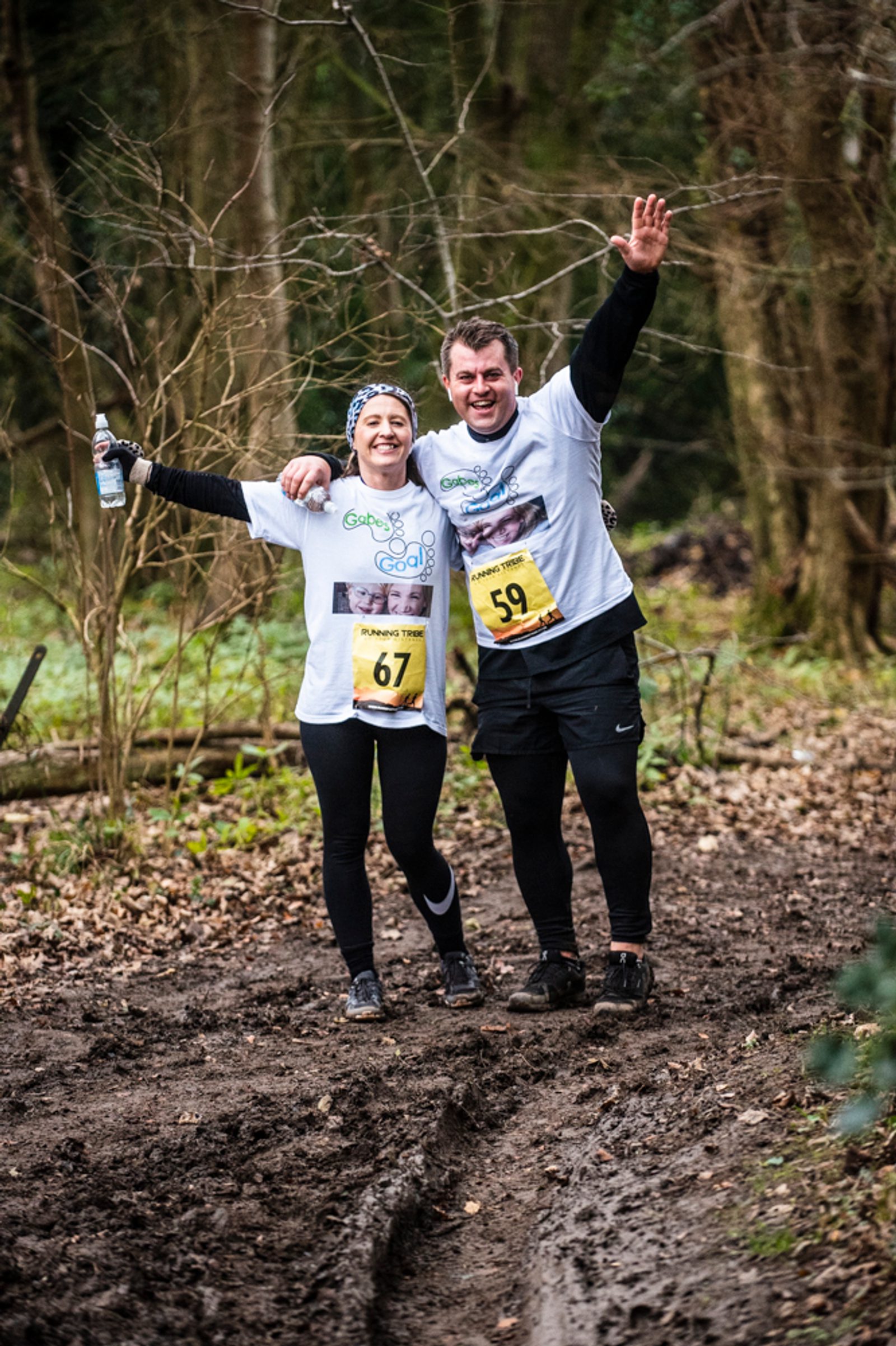 Two smiling runners in white shirts and race bibs (67 and 59) pose together on a muddy forest trail, with one holding a water bottle and the other waving, surrounded by trees and fallen leaves.