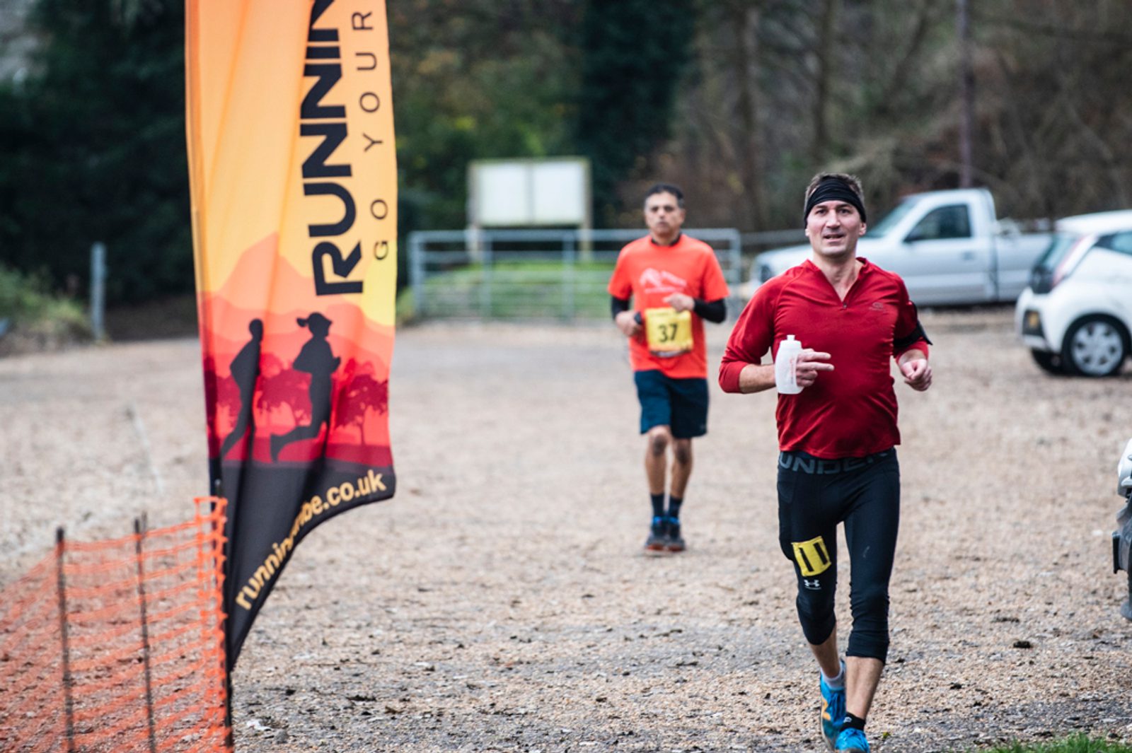 A man in a red shirt and black leggings runs past a "RUNNING" event banner, with another male runner wearing a yellow bib following behind on a gravel path outdoors. Trees and cars are visible in the background.