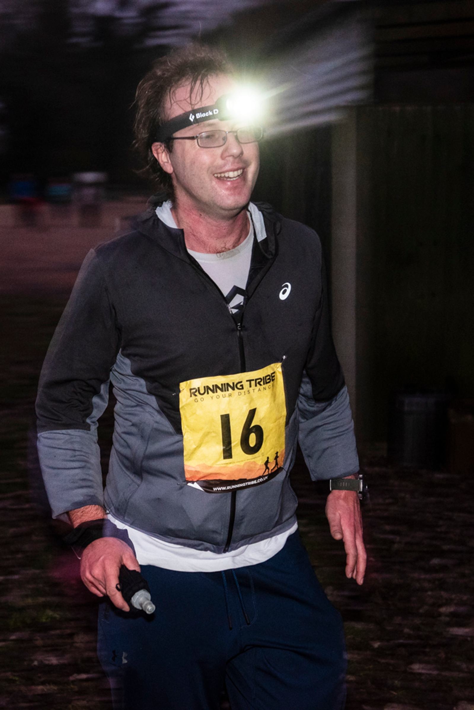 A runner wearing a headlamp and a race number "16" on a yellow bib is jogging outdoors during a night race. He is smiling and holding a small object in one hand. The background is dark with blurred elements.