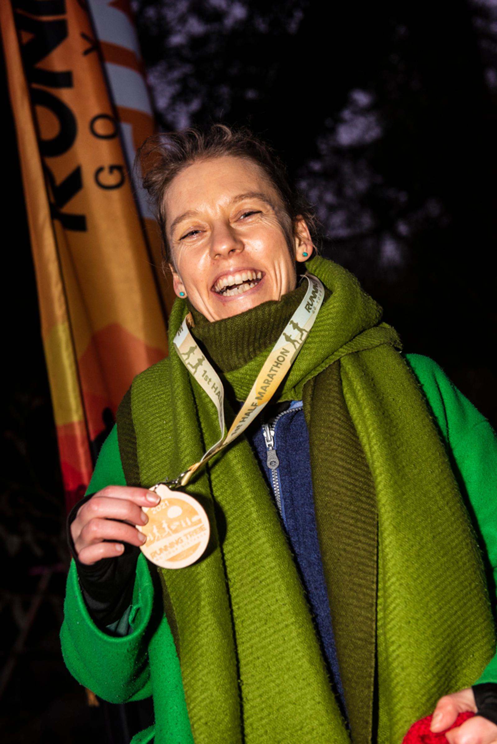 A smiling person wearing a green scarf and a blue jacket holds up a medal. They stand in front of a banner with trees in the background.