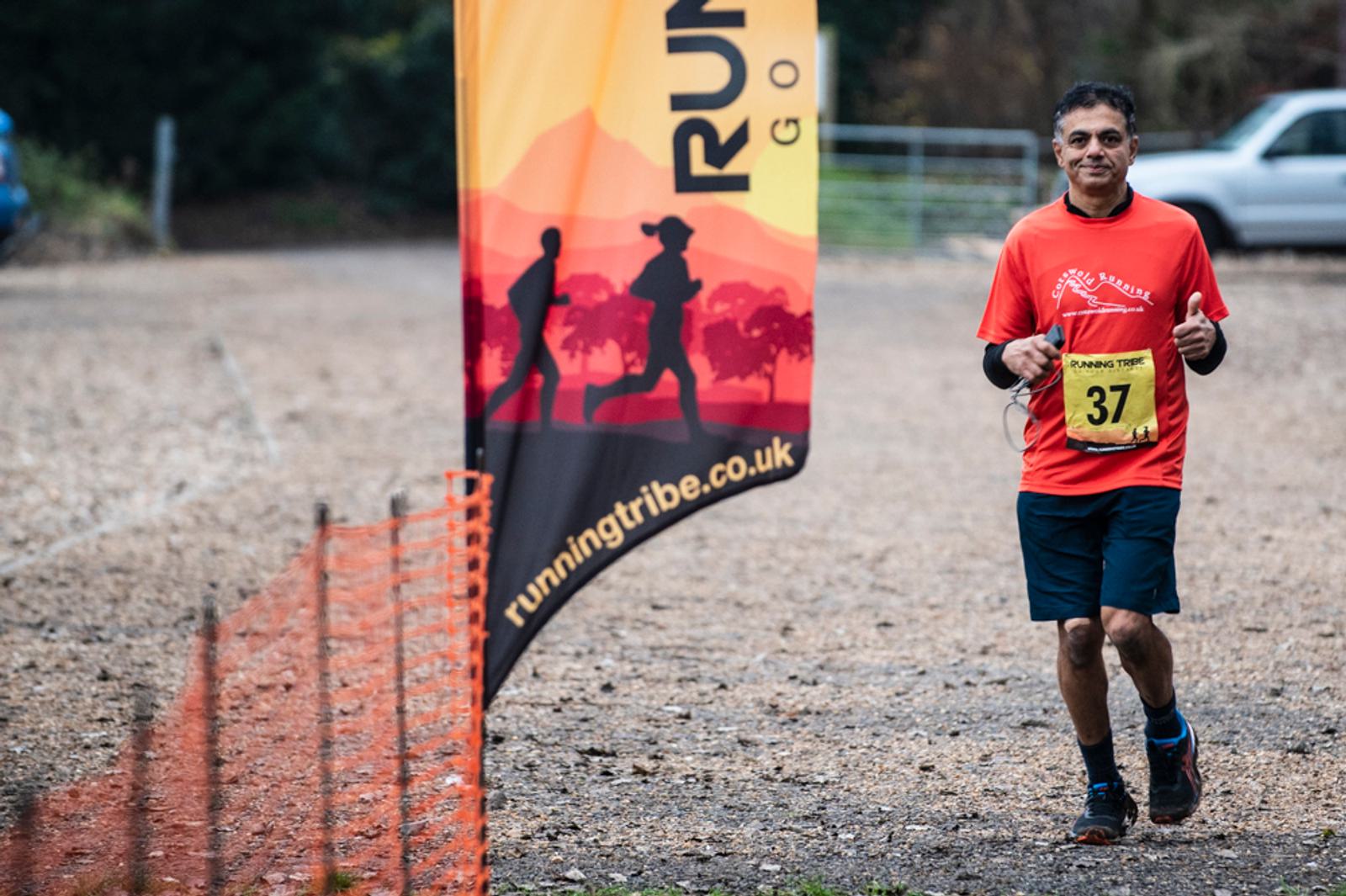 A person wearing a red shirt and black shorts runs on a gravel path, smiling and giving a thumbs-up. They have a yellow race bib numbered 37. A banner with "runningtribe.co.uk" and an image of runners is visible in the background.