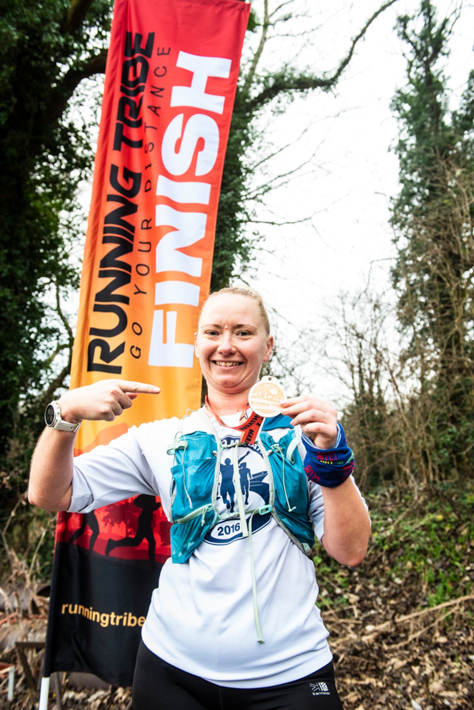 A runner smiles and points at her medal in front of a "Finish" banner. She wears a blue vest and holds the medal up proudly. Trees and bushes are in the background.