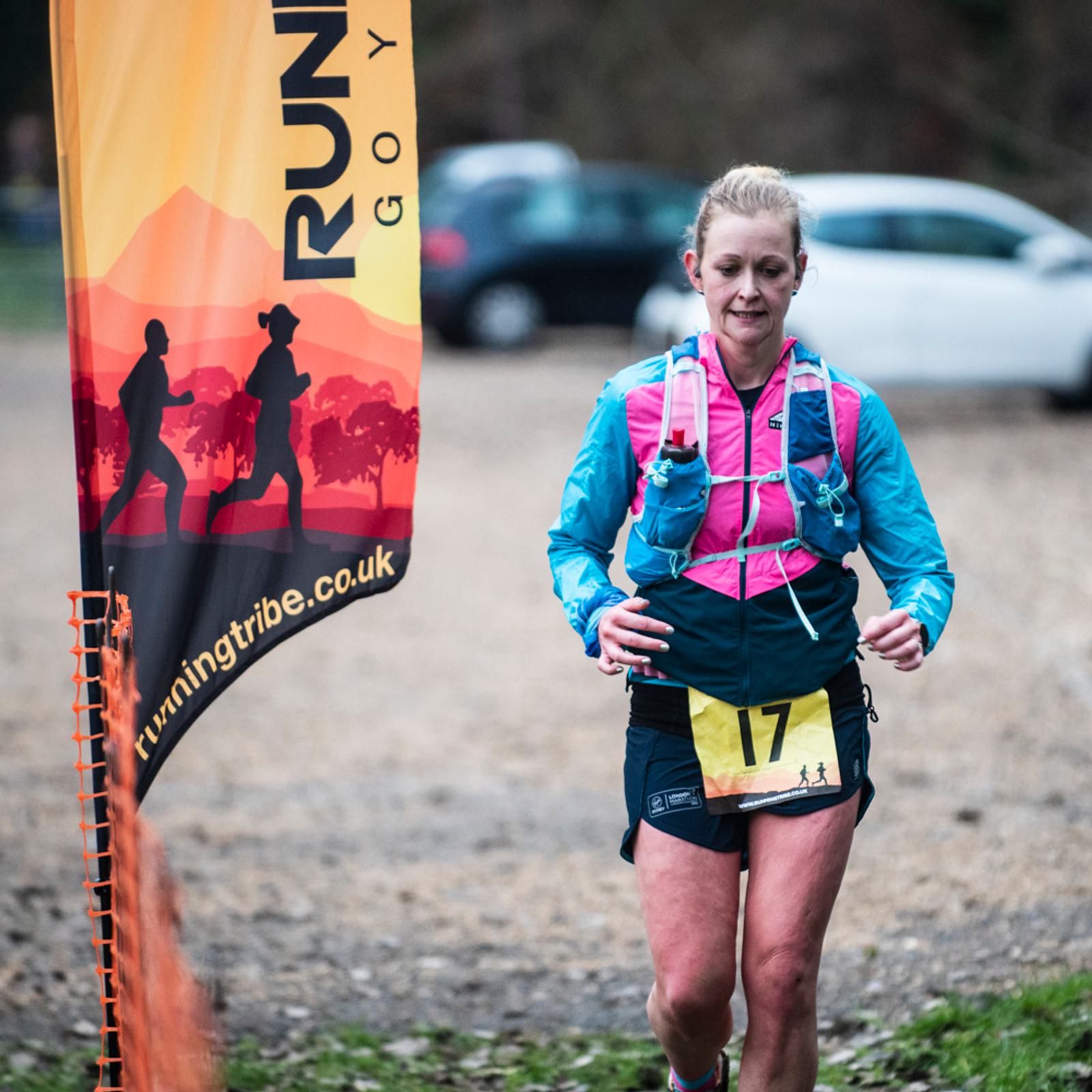 A woman in athletic gear runs past an event flag with silhouettes of runners and the website "runningtribe.co.uk" visible. She wears a race bib numbered 17 and a hydration pack, with parked cars in the background.