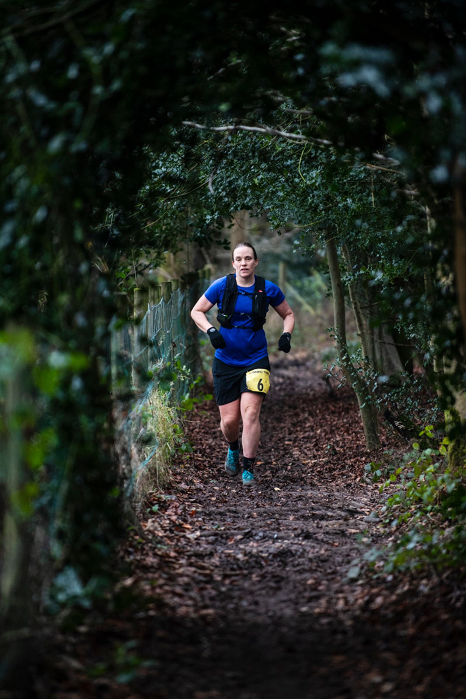 A person wearing a blue shirt and black shorts is running on a narrow, wooded trail. They have a small number bib on their leg. The path is lined with dense green foliage, creating a tunnel-like effect.
