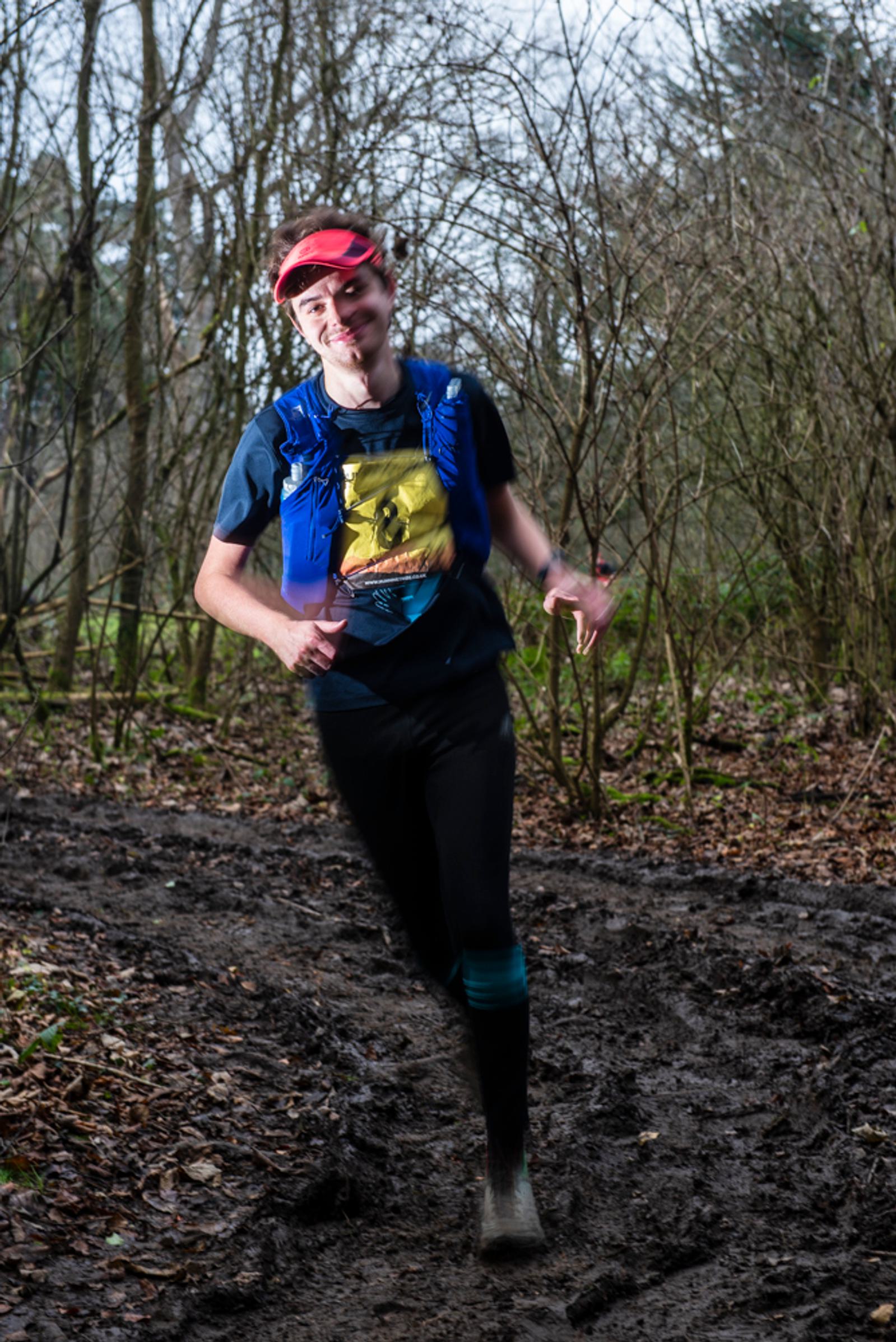A person running on a muddy forest trail, wearing a red cap, a blue hydration vest, and a T-shirt with a graphic print. The trees in the background are bare, suggesting a winter or early spring setting.