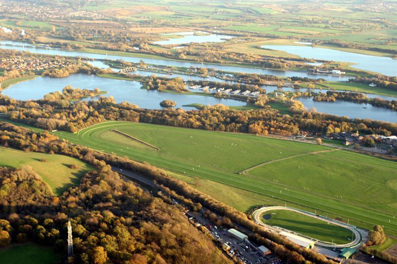 Aerial view of a lush green landscape featuring a large racetrack encircled by a mix of trees and open fields. A river meanders through the background, with nearby buildings and a small marina. Roads and parking areas are visible in the bottom section.