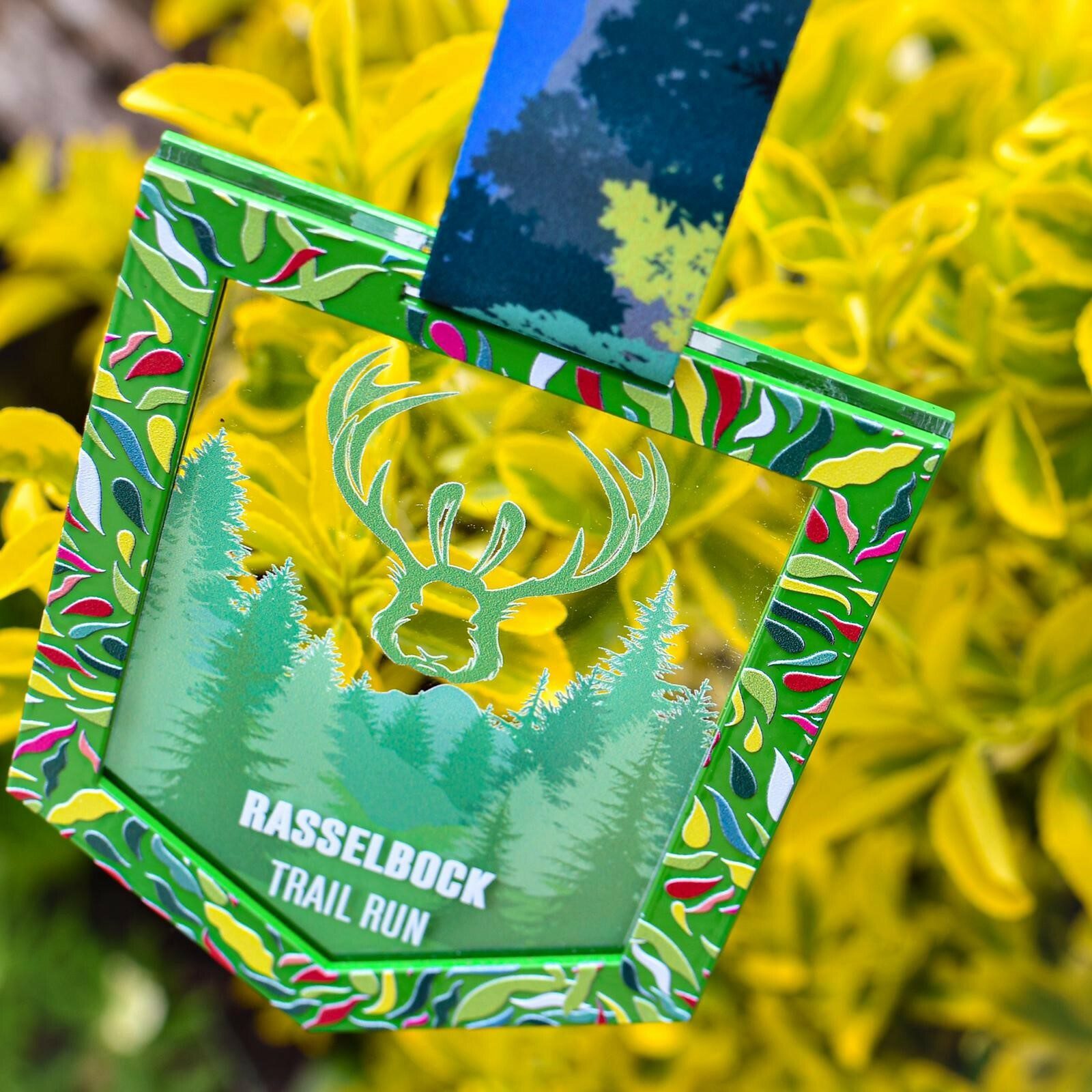 A colorful Rasselbock Trail Run medal with a deer antler design hangs against a backdrop of vibrant yellow foliage. The medal features green and yellow leaf patterns with an illustration of pine trees and a large deer head in the center.
