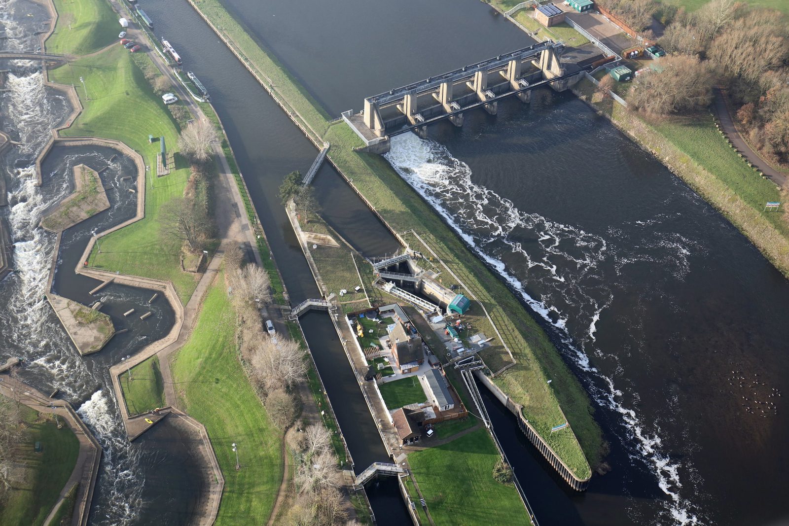 Aerial view of a river with a lock system and dam. The river flows through a series of gates, and adjacent to it, a waterway with multiple lock chambers is visible. Green grassy areas and pathways surround the waterways, and buildings are scattered nearby.
