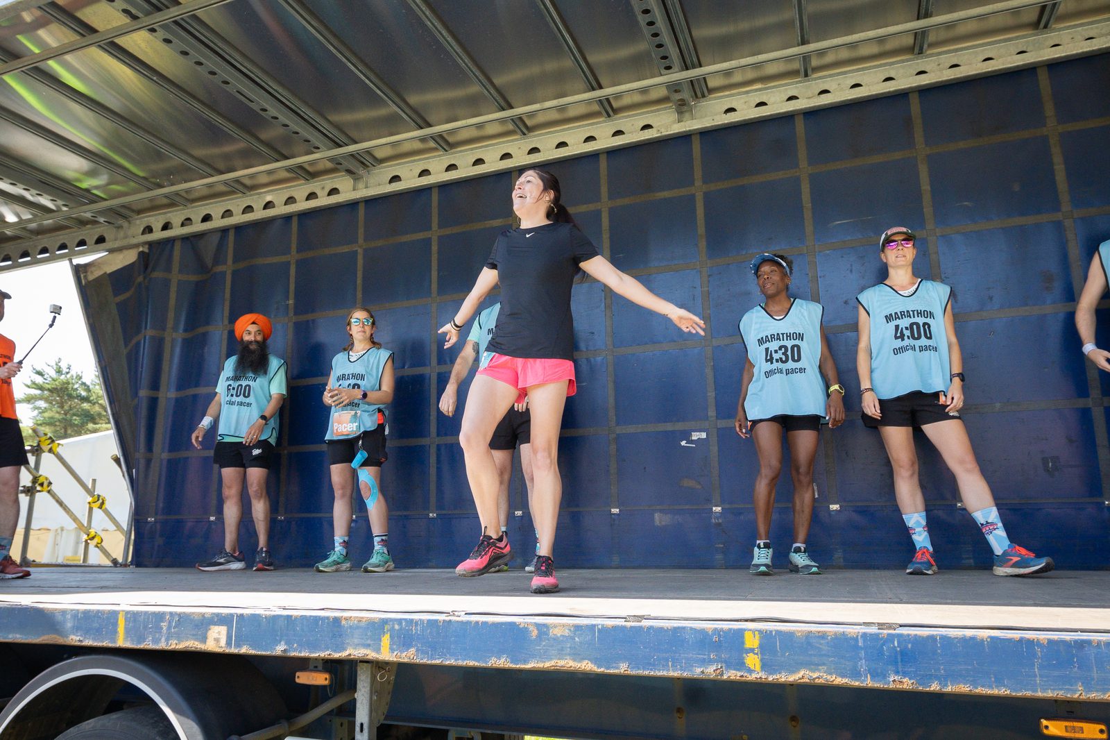 A group of people stand on a stage, with runners in bibs numbered 400, 430, and more. The central figure in a black shirt and pink shorts appears to be leading an exercise or stretching routine. The setting suggests preparation for a running event or marathon.