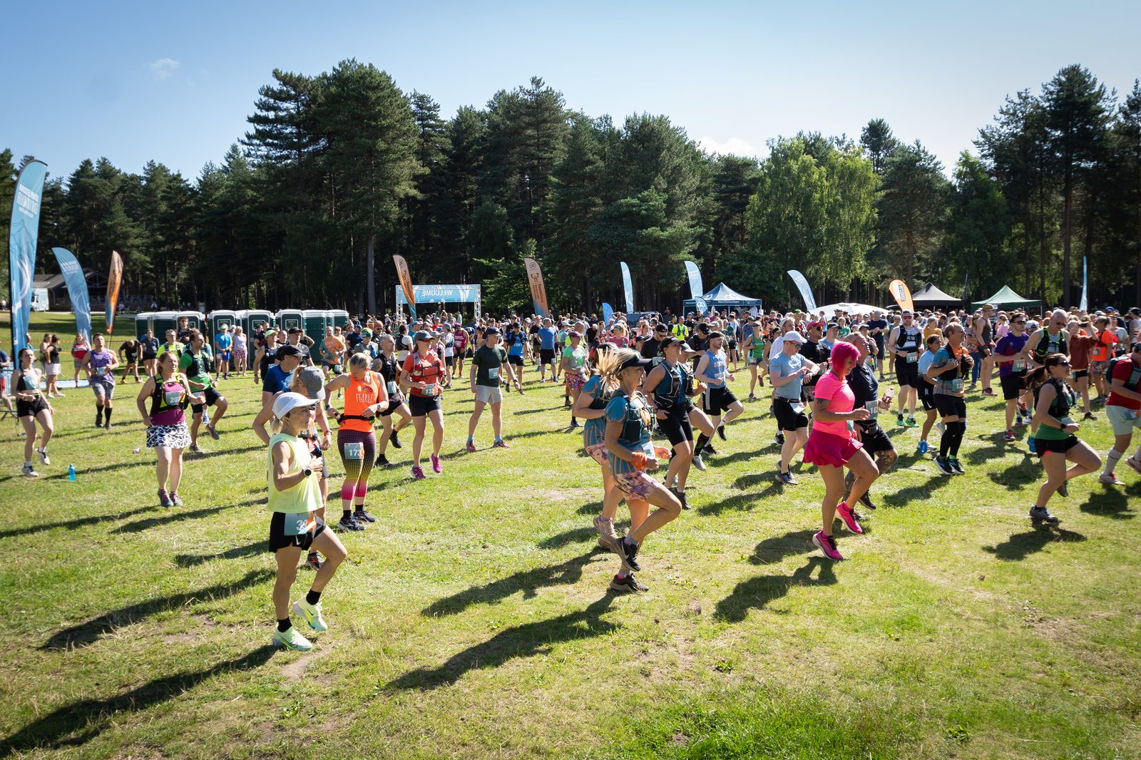 A large group of runners in vibrant athletic attire are participating in an outdoor race on a sunny day. They are running along a grassy field surrounded by trees. Spectators and event tents are visible in the background. Trails of multicolored flags are also seen.