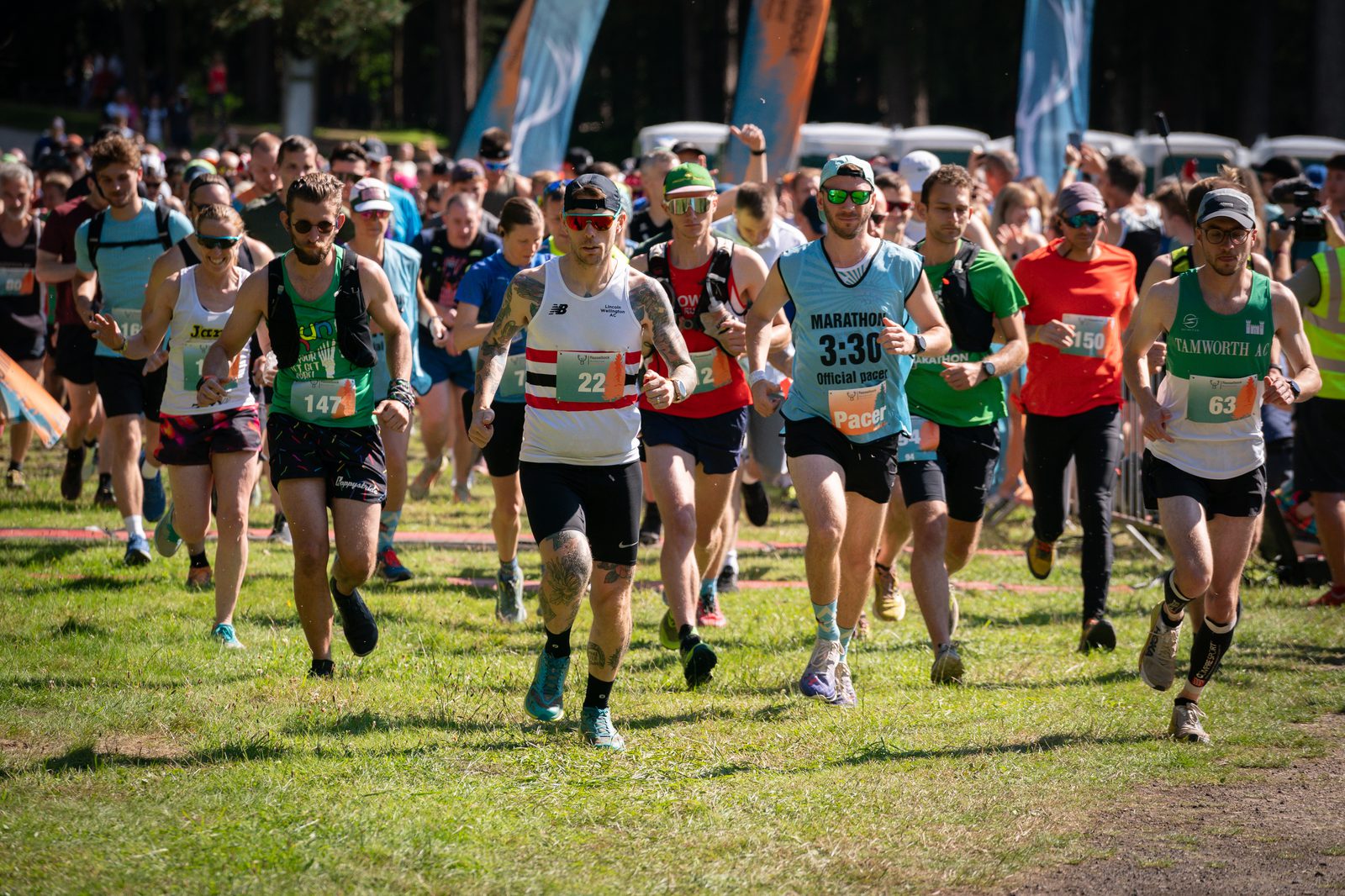 Runners at the starting line of a marathon, with bibs and athletic wear, sprinting forward on a grassy course. A cheerful crowd of participants and supporters is visible behind them, and colorful flags mark the start area on a sunny day.