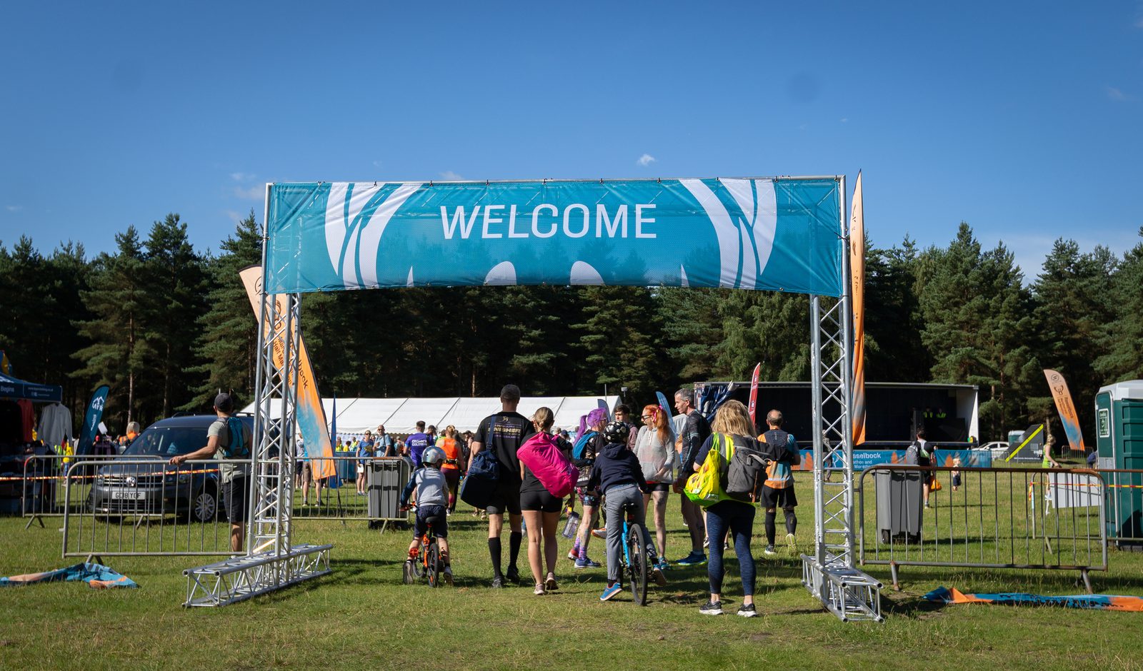 People entering an outdoor event through a large blue and white "Welcome" archway. Several attendees have bicycles and backpacks. The event appears to be in a park, with trees and white tents in the background under a clear blue sky.