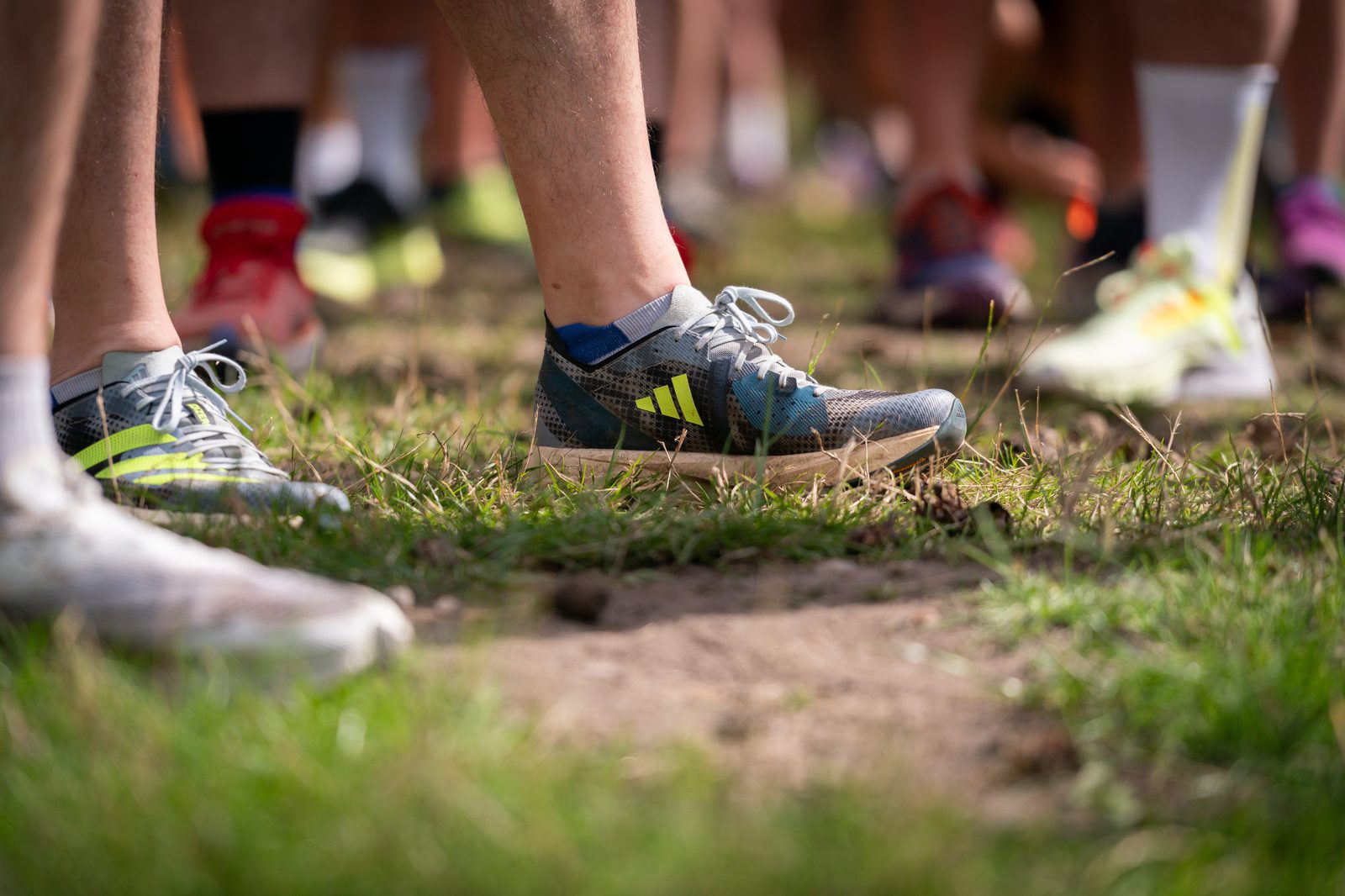 Close-up of runners' feet wearing various athletic shoes standing on a grassy and slightly muddy patch of ground, presumably waiting for the start of a race or event. The focus is on the legs and shoes, with the background blurred.