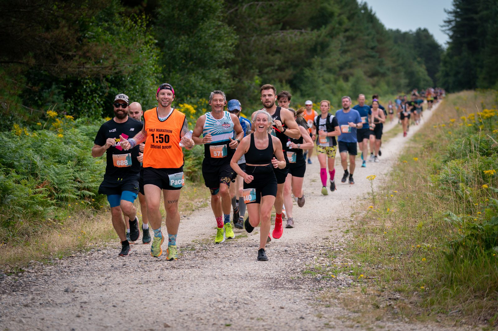 A group of runners participating in a trail running event on a gravel path through a lush, green forest. The runners are at various stages of the race, with some smiling and others focusing intensely. Bright race bibs and clothing add color to the scene.