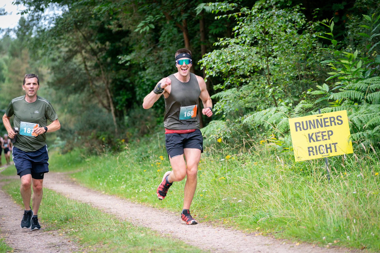 Two runners on a forest trail. The runner in front is wearing a headband, sunglasses, and holding a water bottle, while the second runner follows closely behind. There is a yellow sign on the right side of the trail that reads "RUNNERS KEEP RIGHT.