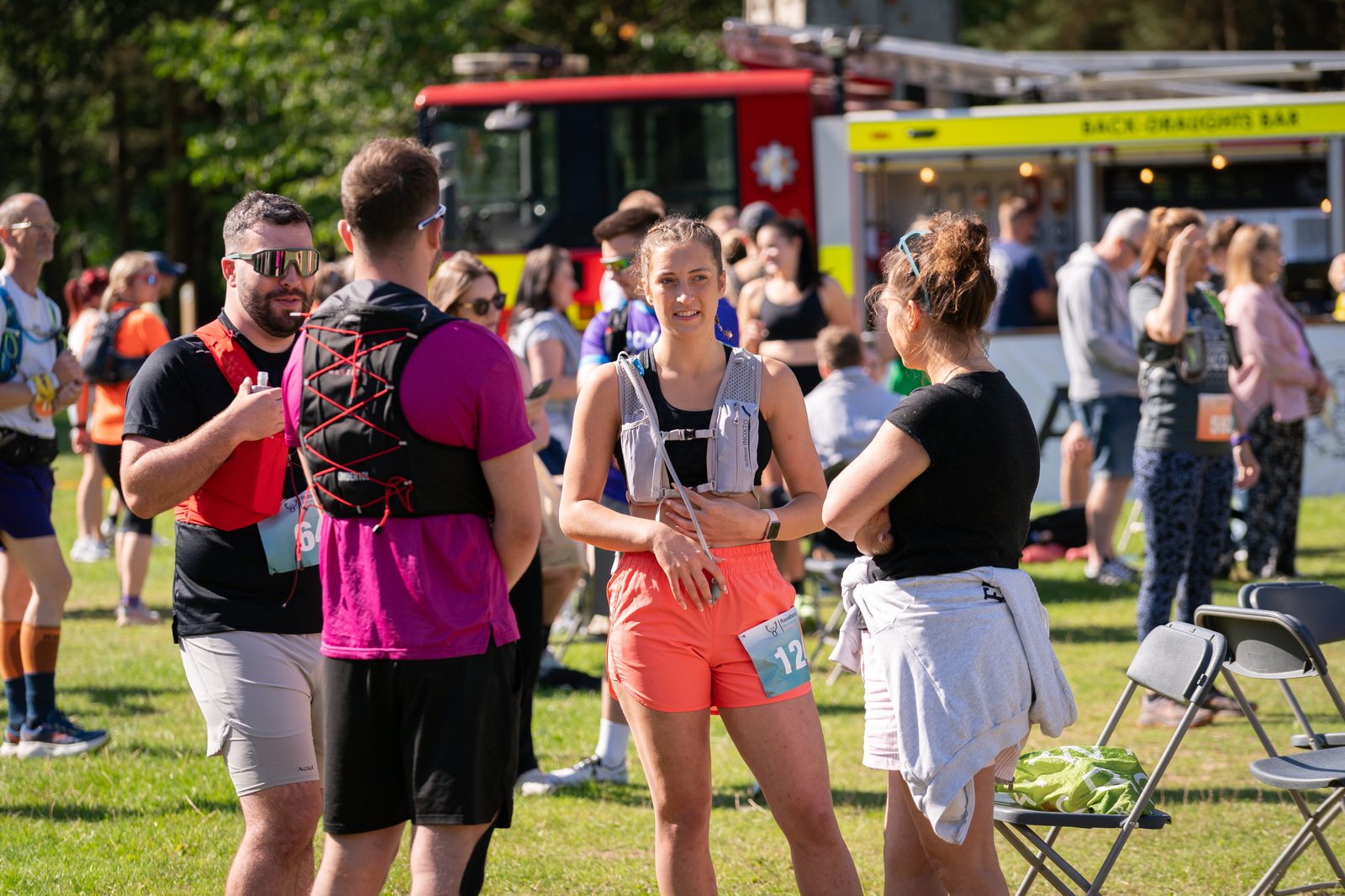 A group of runners gather and chat outdoors on a sunny day. One woman in a gray tank top and orange shorts holds a water bottle. Behind them, other participants and a fire truck are visible. The atmosphere appears lively and social.