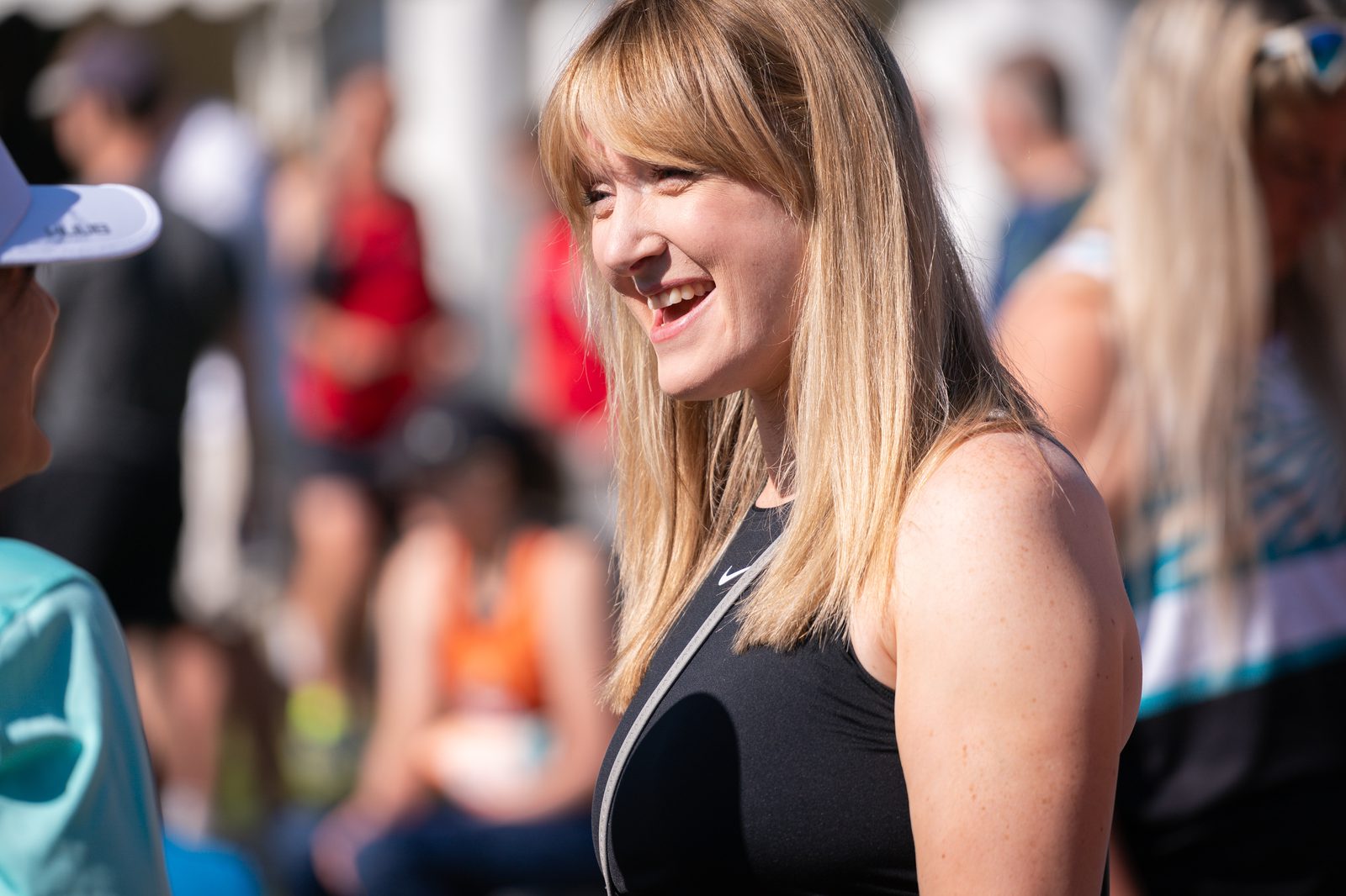 A woman with long blonde hair smiles while talking to a person wearing a cap. She is dressed in a sleeveless black top and is standing outdoors among a crowd of people, some of whom are dressed in athletic attire. The atmosphere appears to be sunny and lively.
