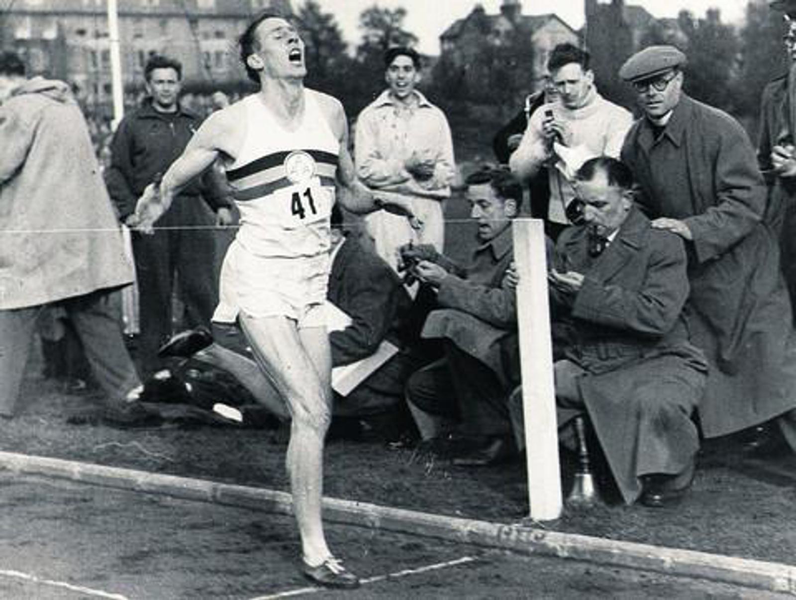 A runner with the number 41 on his bib triumphantly crosses the finish line, appearing exhausted yet victorious. Spectators and journalists, some holding cameras and notebooks, are gathered around to capture the historic moment. The setting appears to be an outdoor track.