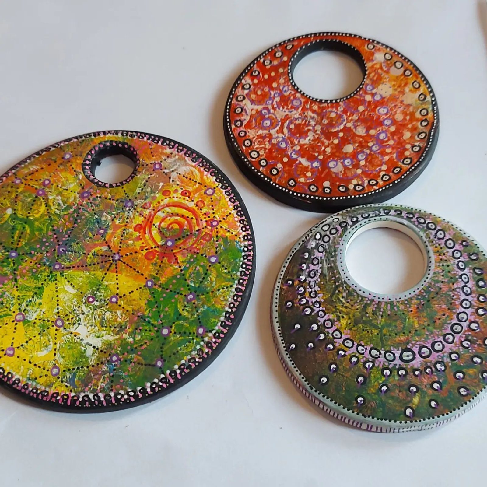 Three intricately decorated circular pendants with colorful designs are set against a white background. The pendants display vibrant patterns of dots and swirls in various hues, including shades of green, yellow, orange, and brown, with small bead-like embellishments.