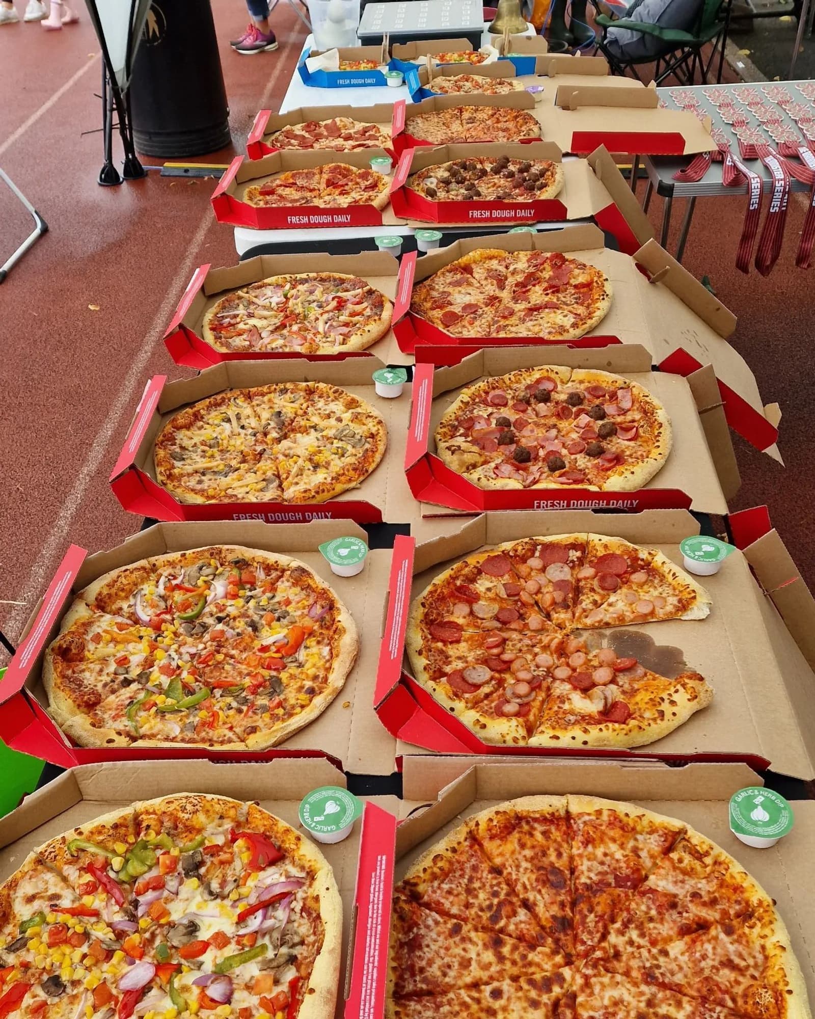 A variety of pizzas are displayed on a table, each in an open box. The pizzas include toppings like pepperoni, mixed vegetables, sausage, and cheese. The table is set on an outdoor track, with people in the background.