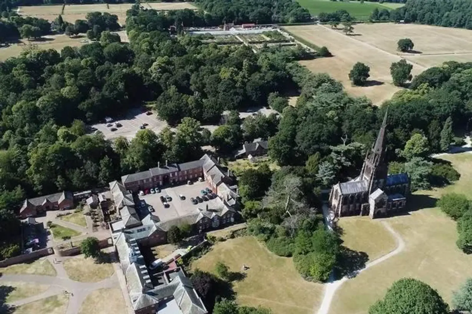 Aerial view of a countryside estate featuring a historic church with a tall spire, surrounded by lush green trees and grass. Adjacent buildings are scattered throughout the landscape, with a distant parking area and fields visible.