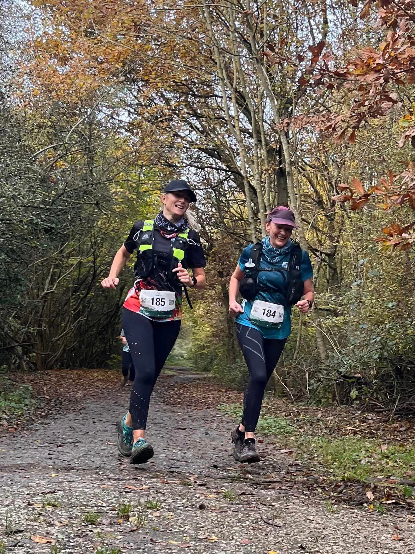 Two people are running on a forest trail with autumn leaves scattered around. Both wear numbered race bibs and athletic gear. They appear cheerful as they jog side by side under a canopy of trees.