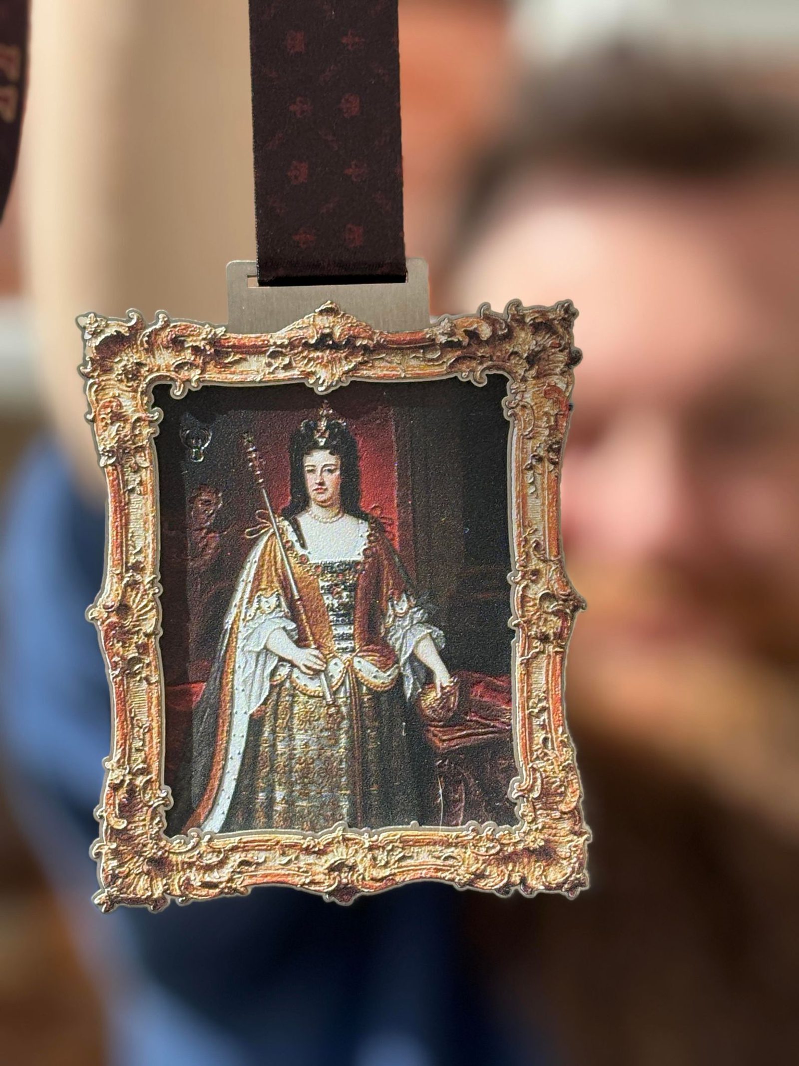 A close-up view of a person holding a decorative lanyard with a detailed, ornate frame. The frame encases an image of a regal figure adorned in royal attire, holding a scepter. The background is slightly blurred, and the person's face is intentionally out of focus.