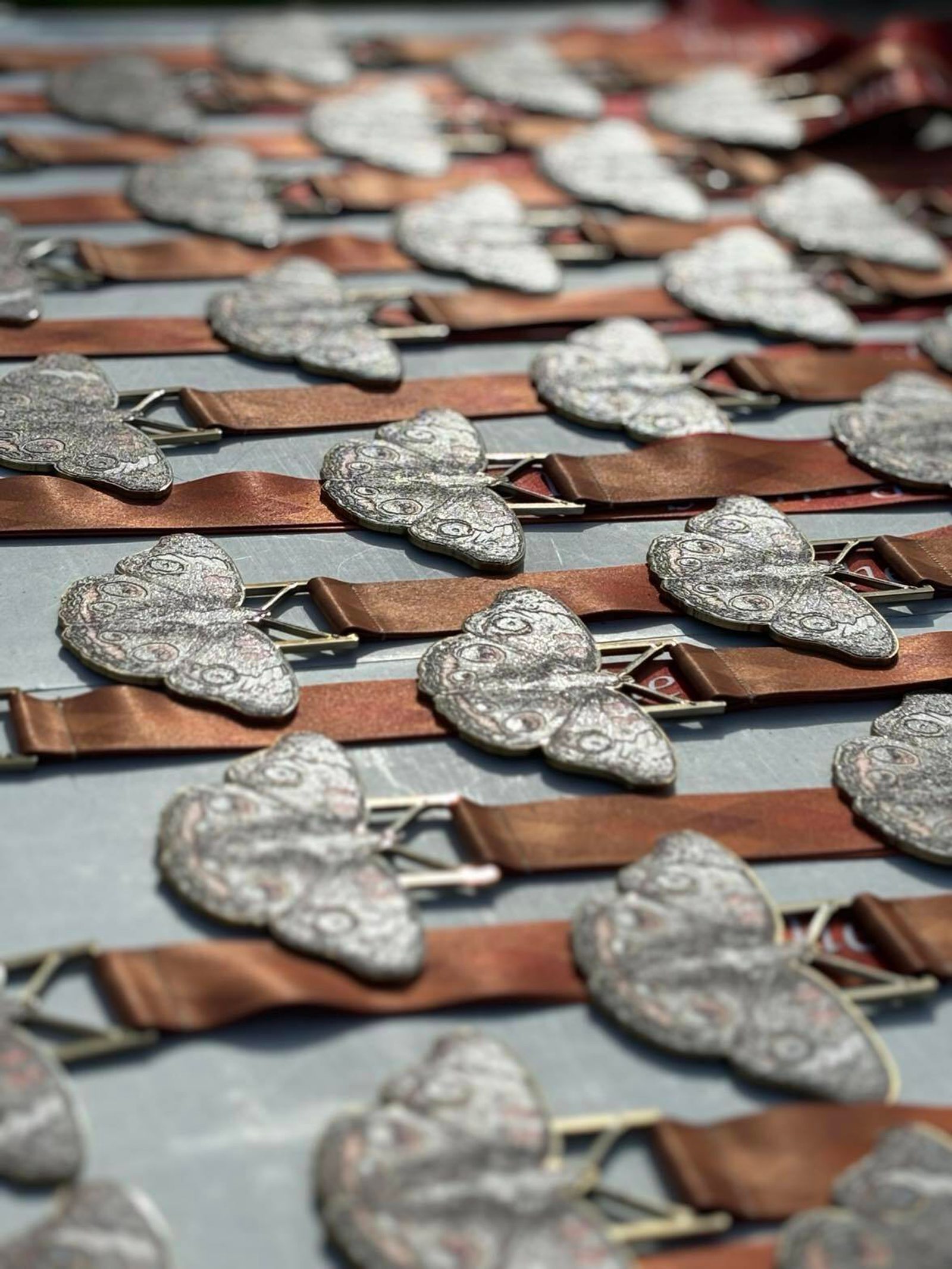A close-up of decorative silver buckles attached to brown straps, arranged in neat rows on a flat surface. The buckles have intricate designs and are arranged in a visually appealing, repetitive pattern.
