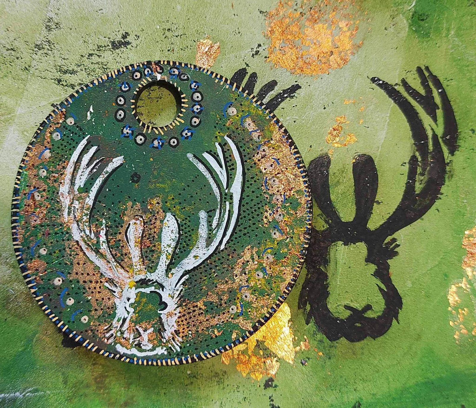 A piece of mixed media art featuring a circular green disc with a white, abstract deer head design in the center, adorned with small decorative elements. In the background, there is a black silhouette of a deer’s head against a textured, green and yellow backdrop.
