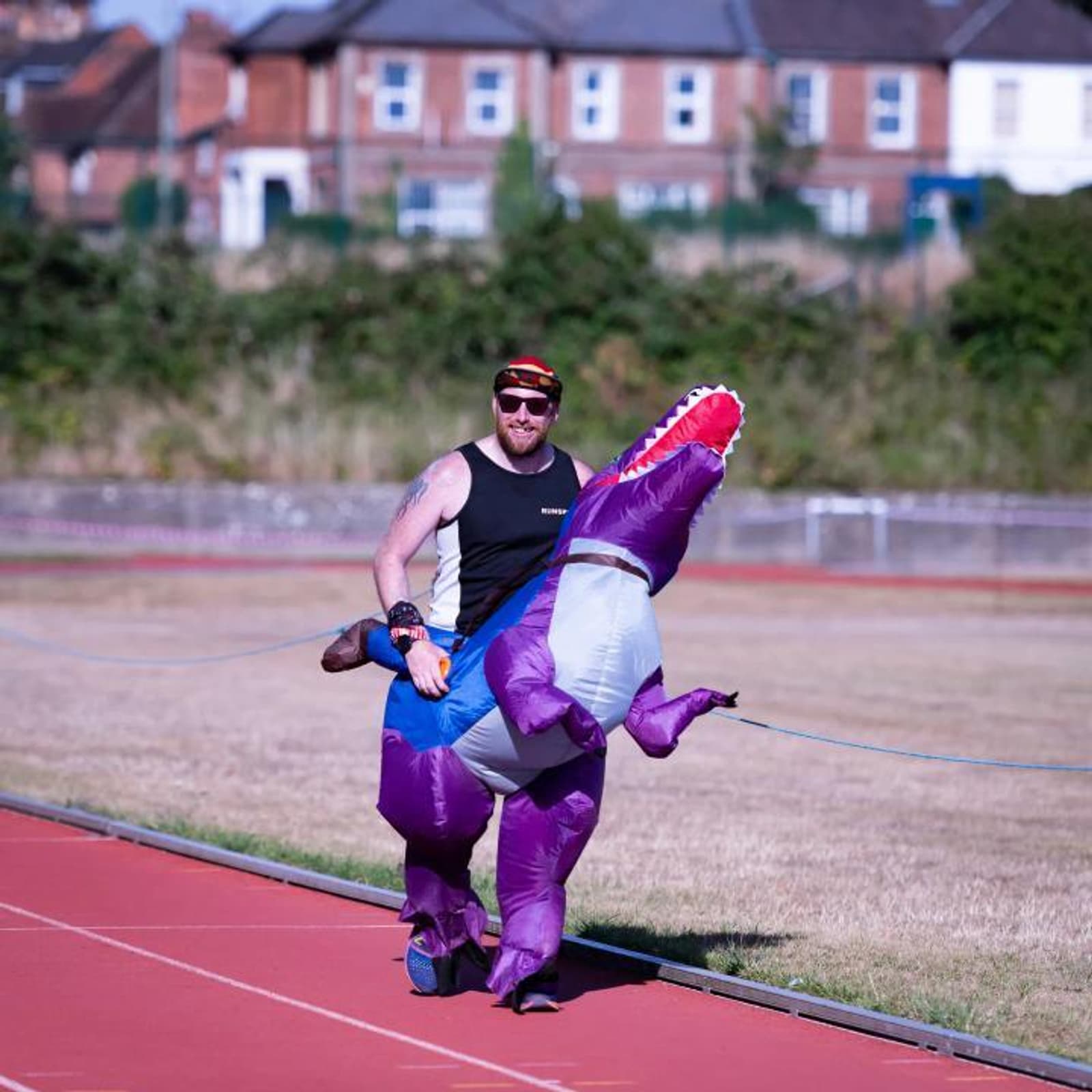 A person wearing a colorful inflatable dinosaur costume jogs on a red track. They are outdoors on a sunny day with a background of houses and greenery. The person is also wearing sunglasses and a headband.