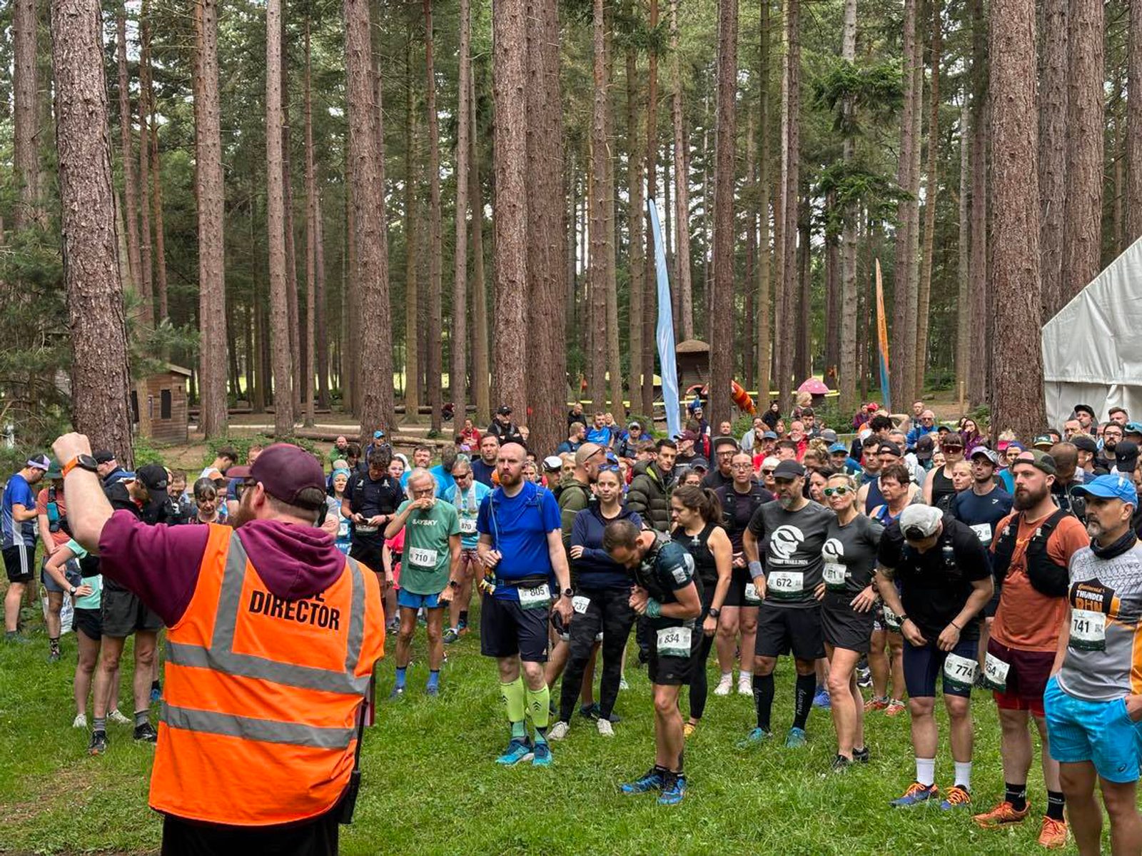 A person wearing an orange vest with "Director" written on the back addresses a large group of runners gathered at a start line in a forest. The runners are wearing bibs with numbers and appear ready to start a race. Tall trees surround the clearing.