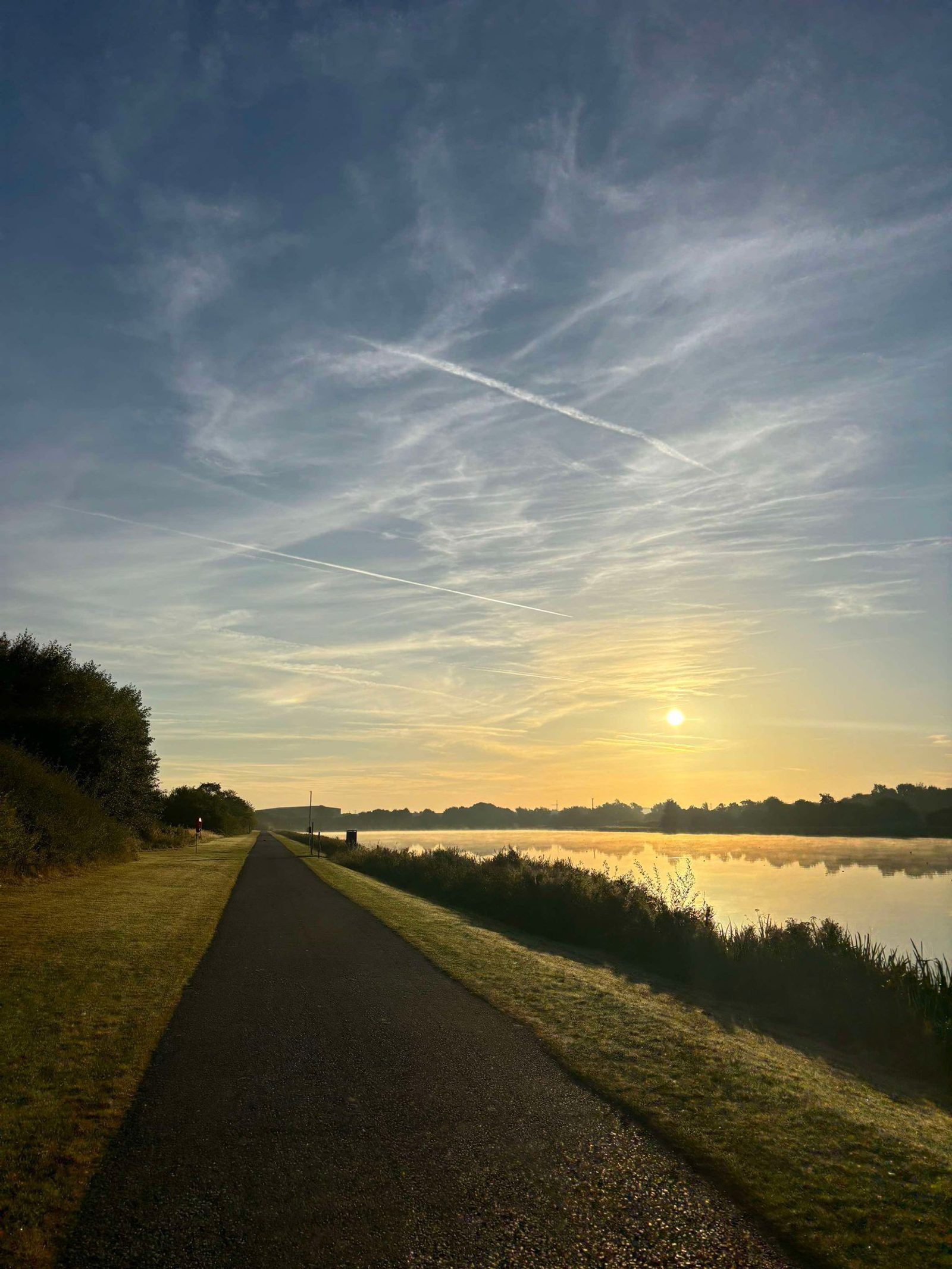 The image shows a serene riverside path at sunrise. The sun is low on the horizon, casting a golden hue over the landscape. The sky is dotted with thin, wispy clouds, and the calm river mirrors the morning light. Dense foliage lines one side of the path.