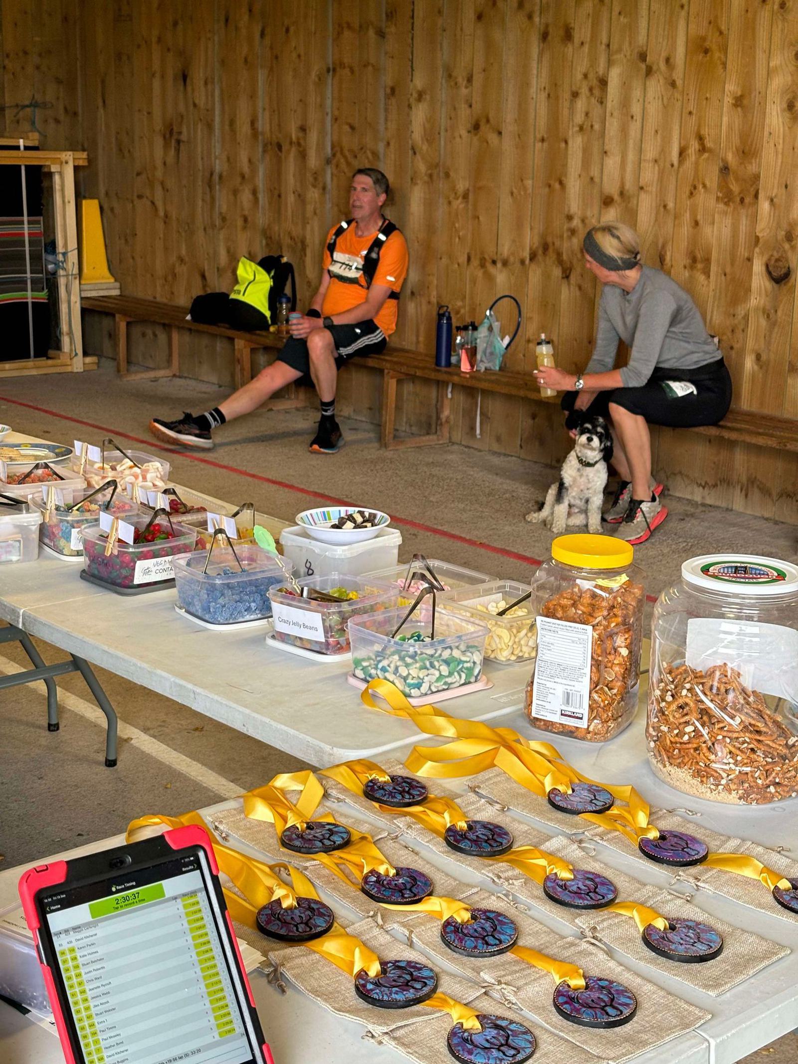A man and a woman sit on a bench in a wooden room. The man is wearing cycling gear, and the woman is with a small dog. A table in the foreground displays medals, snacks, and a tablet with a list.