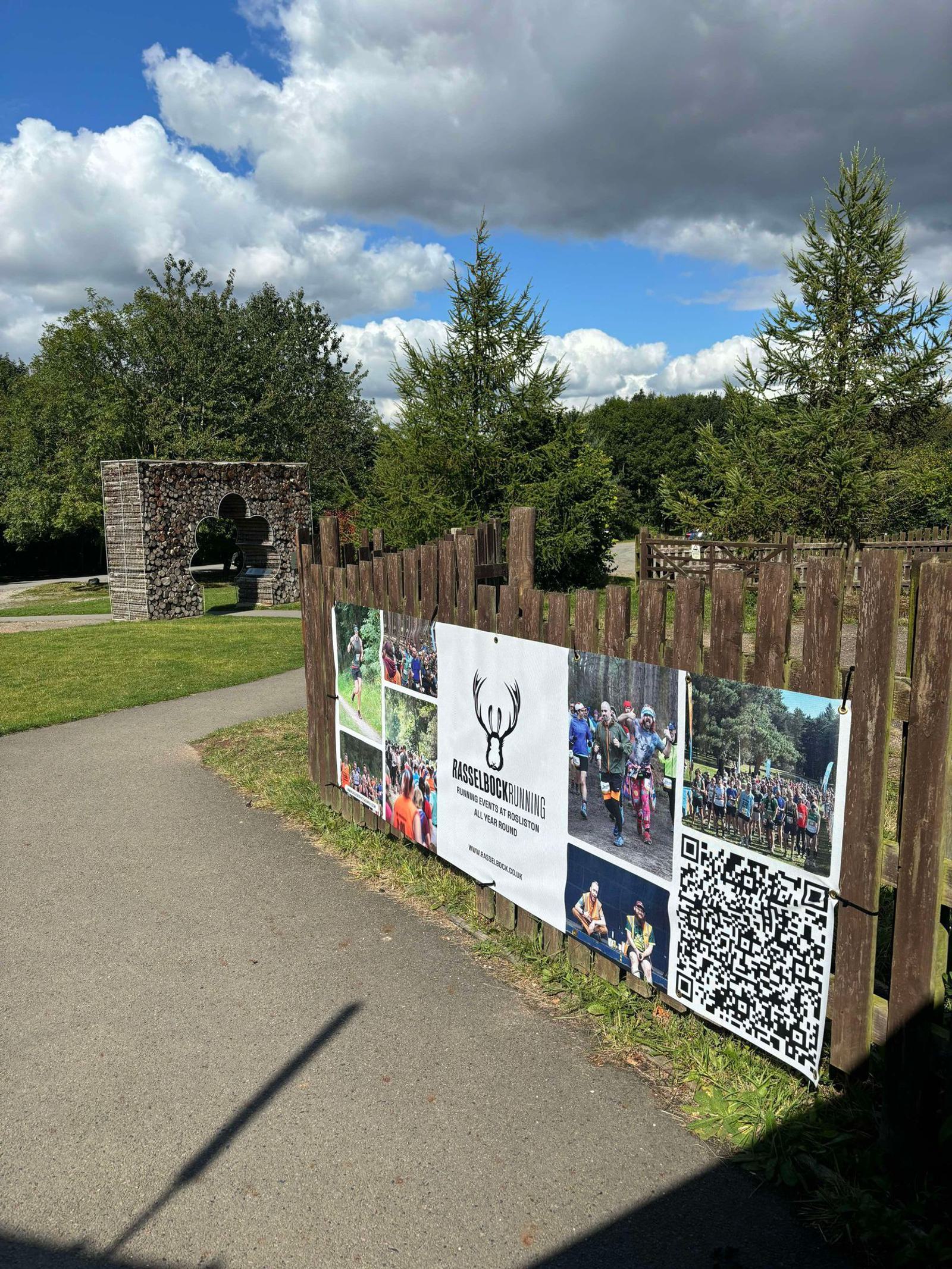 A wooden fence displays a banner with photos of a running event and a QR code. The background shows a grassy area, a stone arch, and several trees under a partly cloudy sky.
