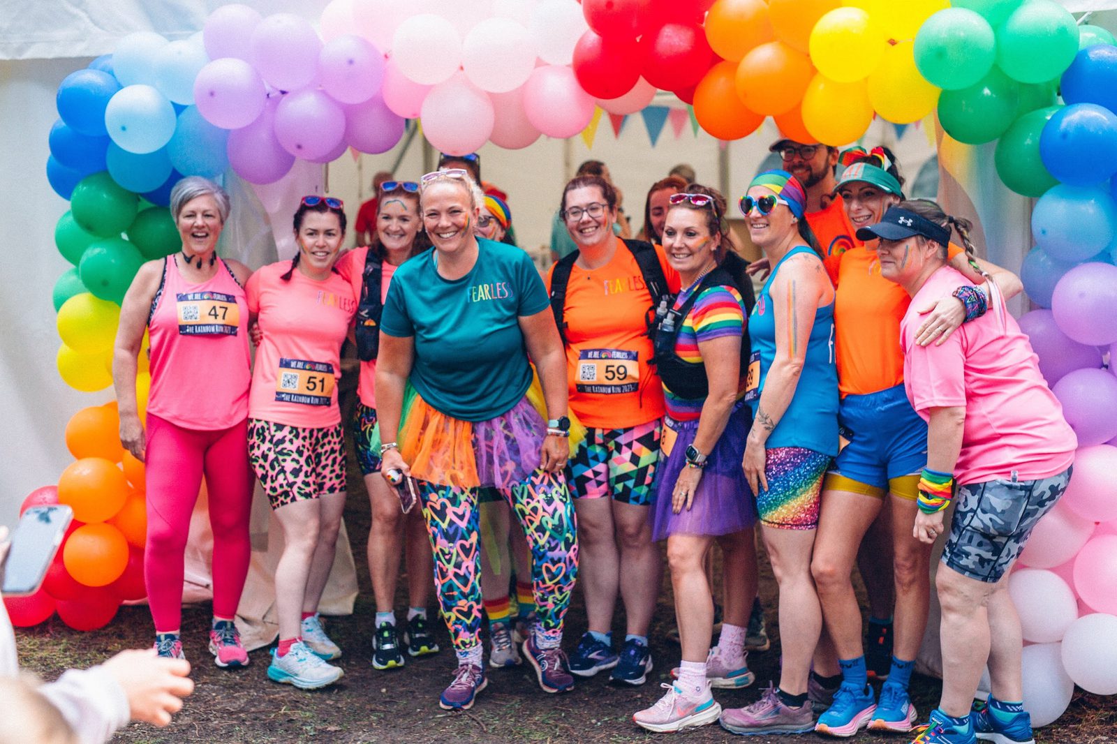 A group of people dressed in vibrant, colorful athletic wear pose together under a rainbow balloon arch. They appear to be participating in a festive fun run or event, with numbers pinned to their shirts and joyful expressions on their faces.