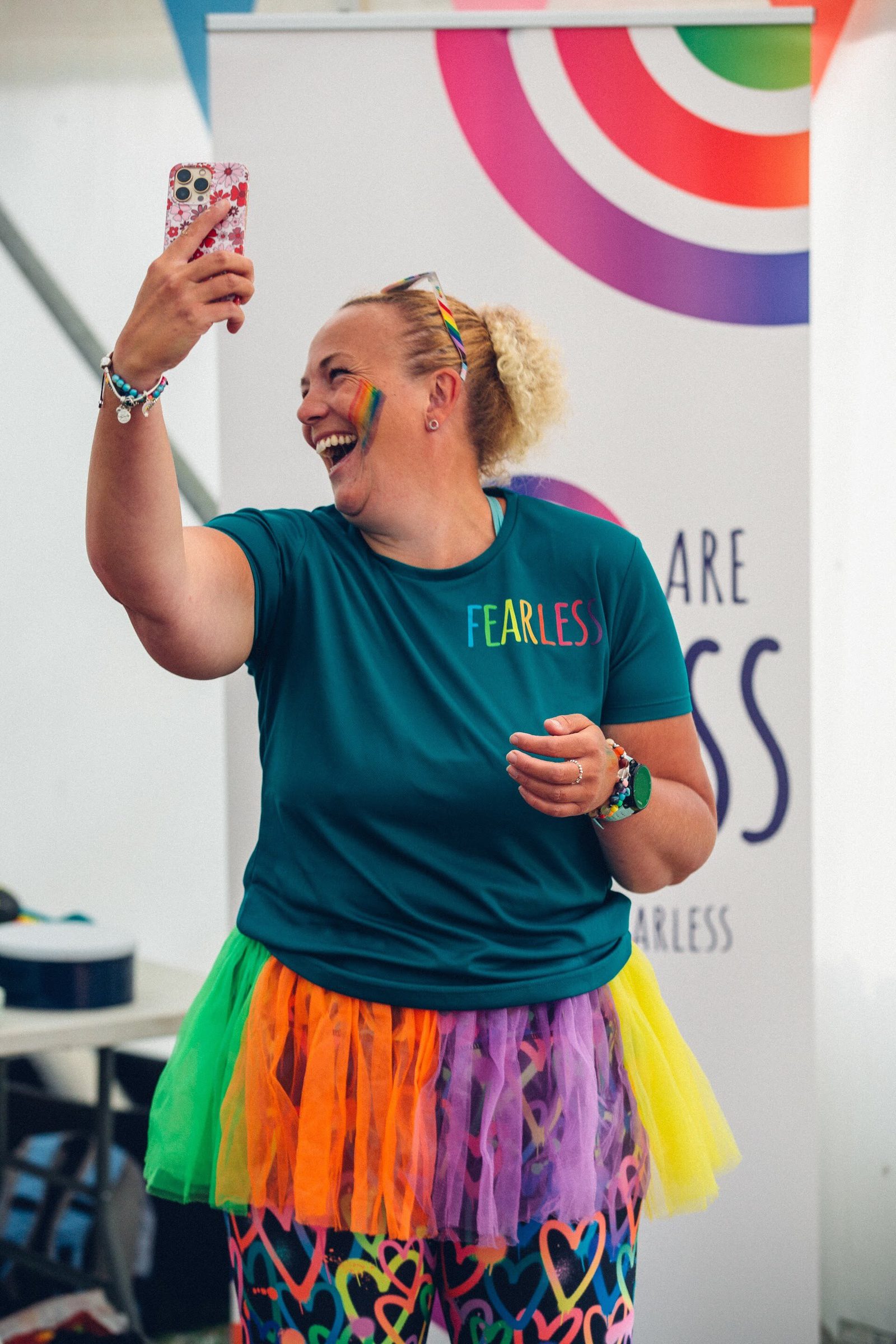 A person with curly hair is smiling and holding up a smartphone to take a selfie. They are wearing a colorful tutu, a 'FEARLESS' t-shirt, and have rainbow face paint. There is a banner with colorful graphics in the background.