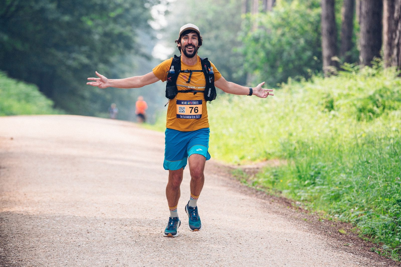 A smiling man wearing a yellow shirt, blue shorts, a cap, and a hydration pack is running joyfully on a forest trail. His bib number is 76 and his arms are outstretched. The background includes another runner in the distance and a pathway bordered by greenery.