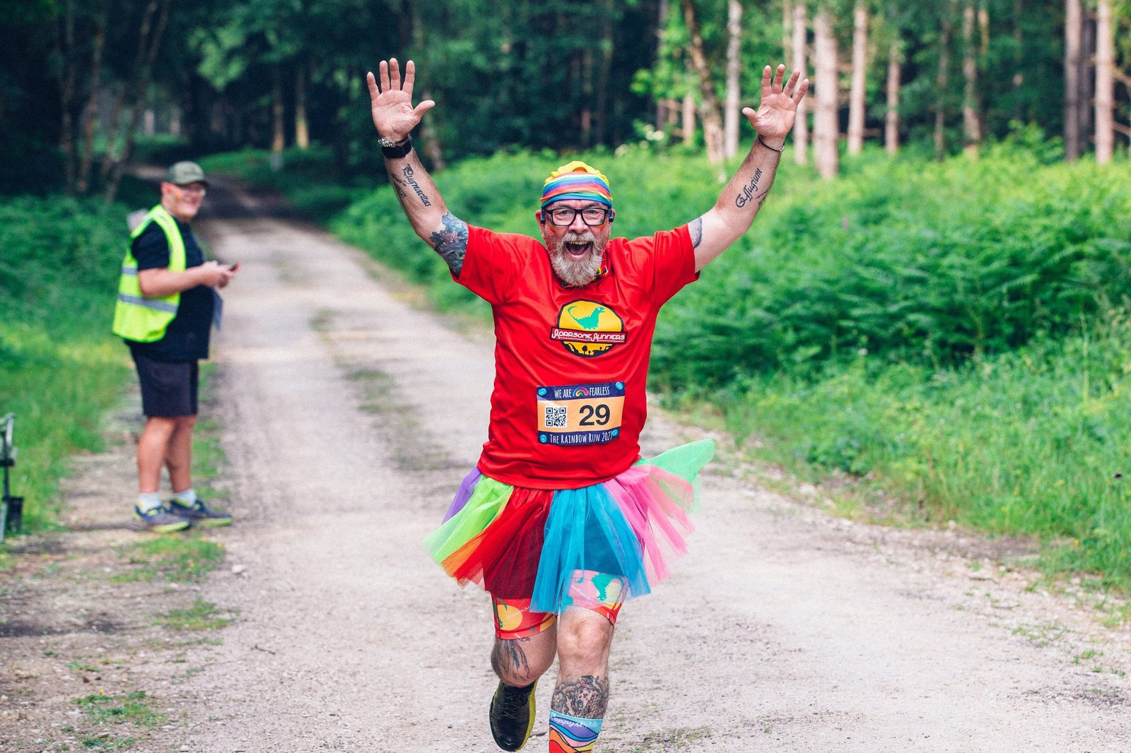 A person with a beard and colorful headband is joyfully running on a forest path during a race. They are wearing a red shirt, rainbow tutu, and leggings, with both arms raised in excitement. Another person in the background is capturing the moment.