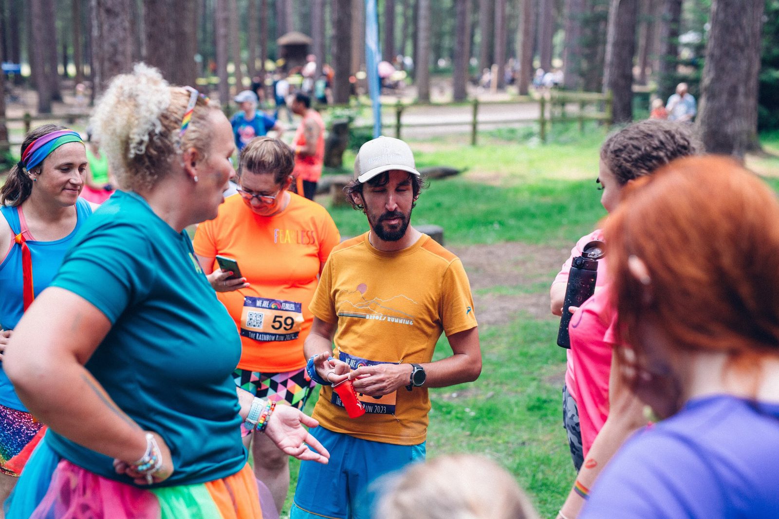 A group of people are gathered in a forested area, some wearing colorful athletic clothing and race bibs. One person in a yellow shirt and cap appears to be giving instructions or speaking to the group. Trees and other participants are visible in the background.