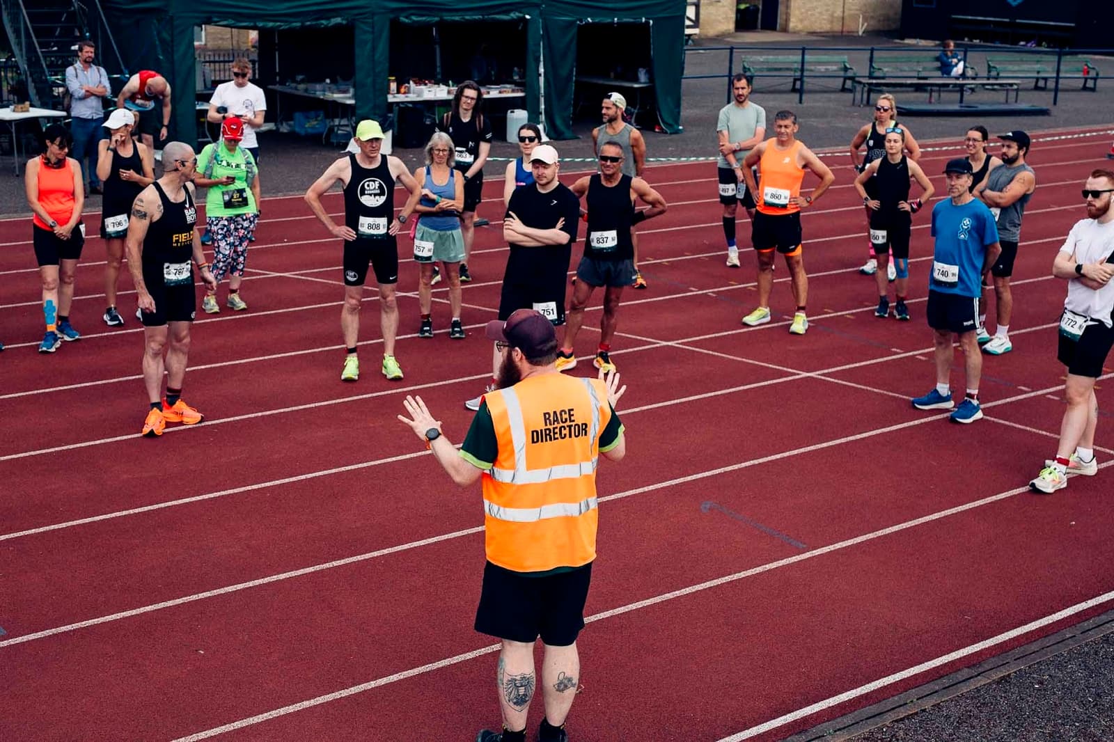 A group of runners stands on a track listening to a person wearing a "Race Director" vest. The runners wear athletic gear and numbered bibs. In the background, there's a green tent and tables.
