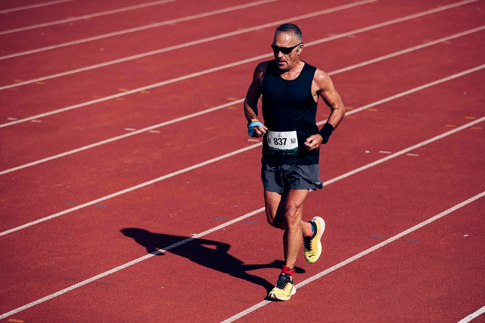 A person wearing sunglasses, a black tank top, and shorts runs on a red track. They hold a small water bottle and wear a race bib numbered 837. The sun casts a shadow on the track.