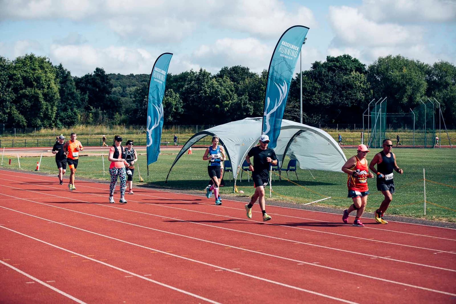 People are running on a red track with a tent and blue flags in the background. The sky is partly cloudy and there are trees in the distance. Some individuals are wearing colorful athletic gear.
